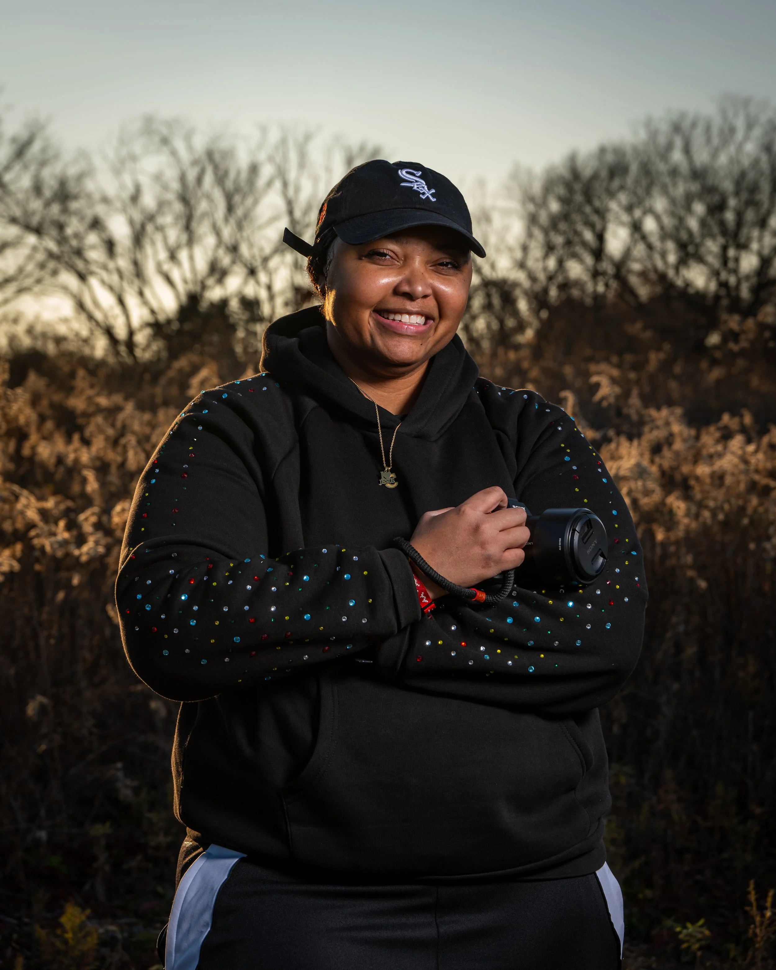 A woman smiling outdoors during sunset, wearing a black hoodie with colorful dots, a black cap, and holding a camera.