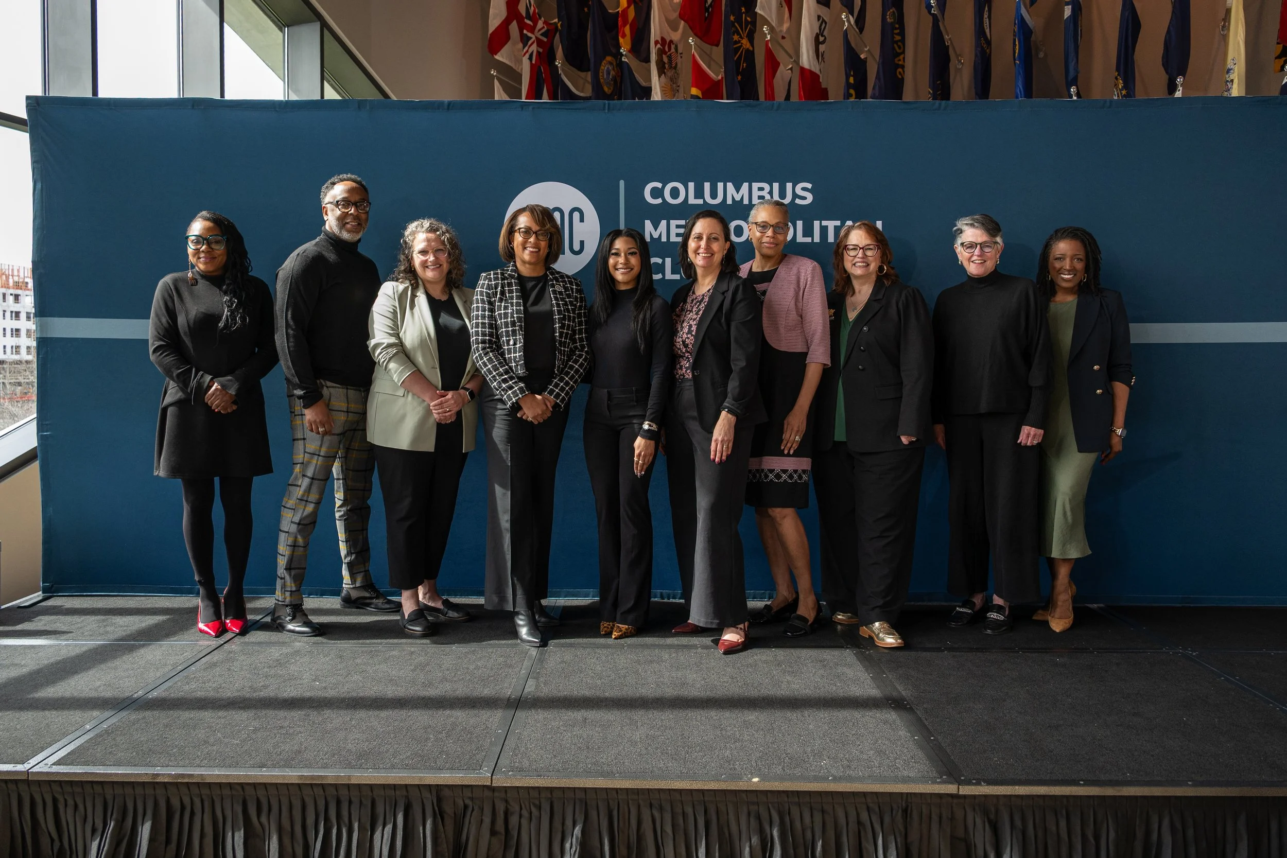 Group of ten women standing side by side on stage in front of a blue backdrop with a logo and text, at an event for the Columbus Metropolitan Library, with flags hanging above in a large indoor space.