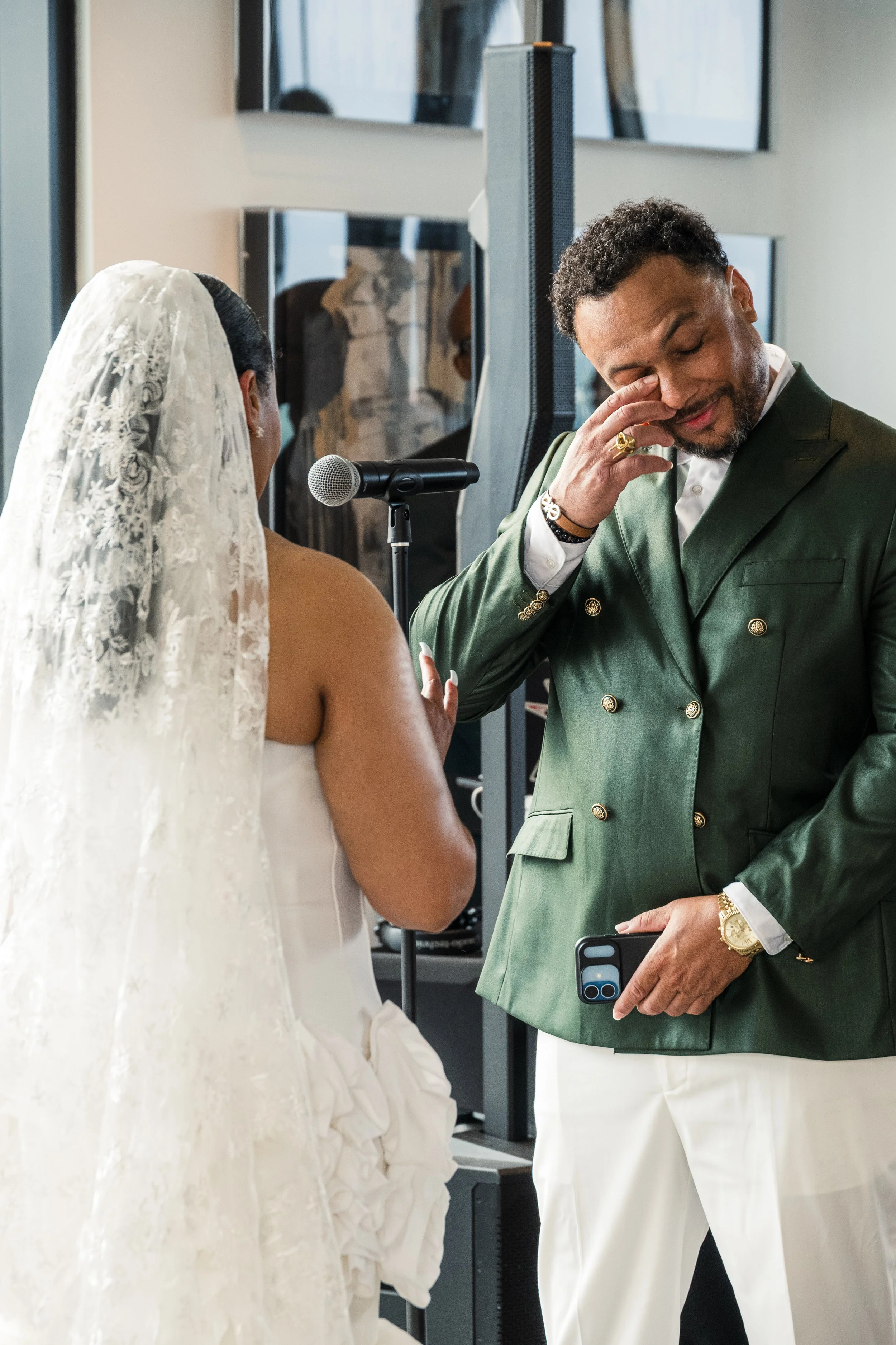 A bride and groom during their wedding ceremony, with the groom wiping a tear from his eye, standing next to a microphone, in a bright indoor setting.