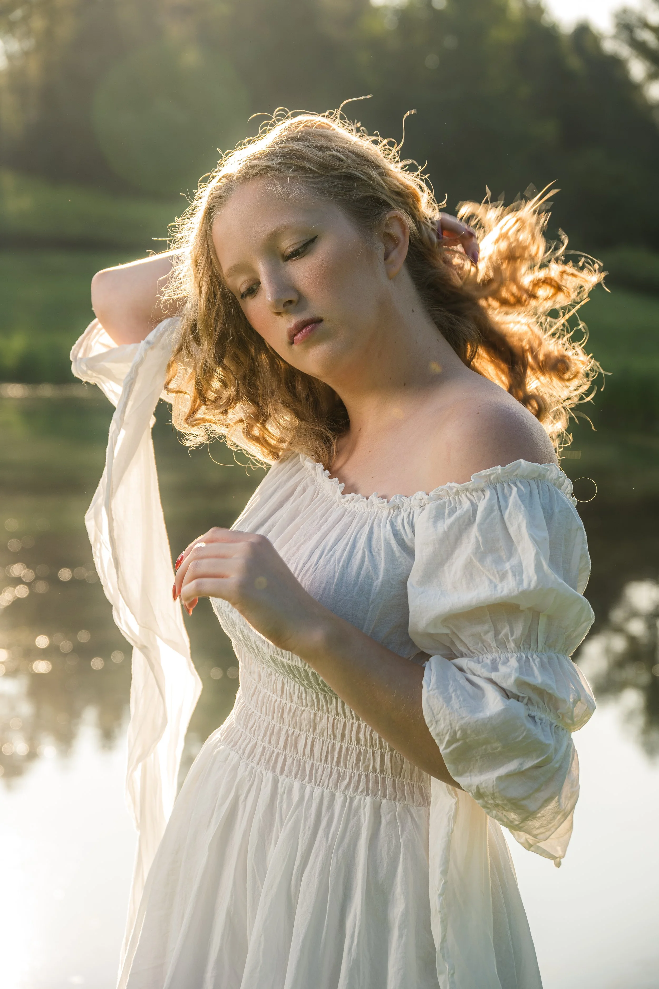 Young woman in a white dress standing outdoors near a body of water with sunlight shining behind her, with her eyes closed and hand raised to her head.