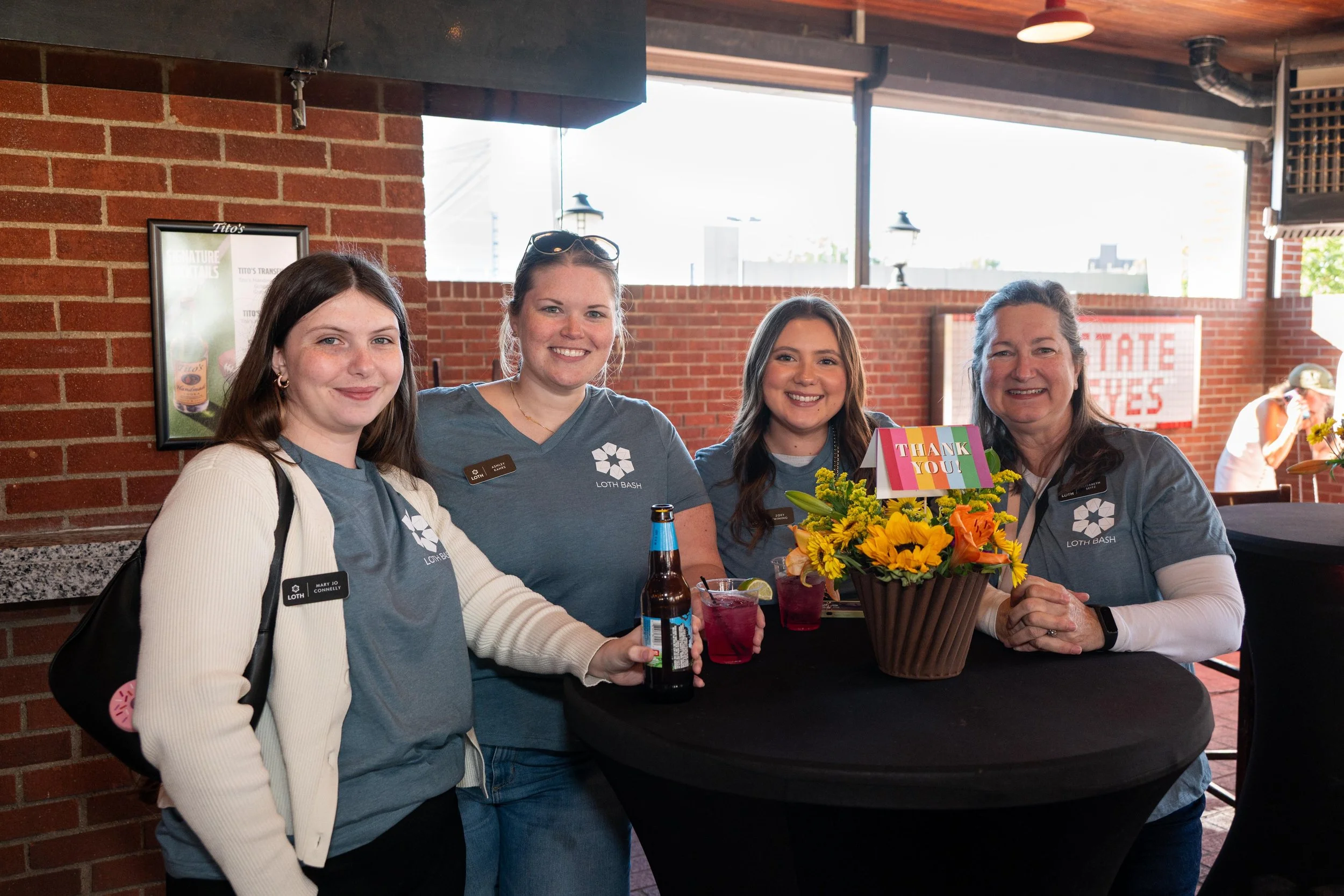 Four women smiling at a table decorated with a colorful flower arrangement and a "Thank You" sign. The women are wearing gray T-shirts with a logo and name tags, standing against a brick wall with windows and outdoor scenery visible.