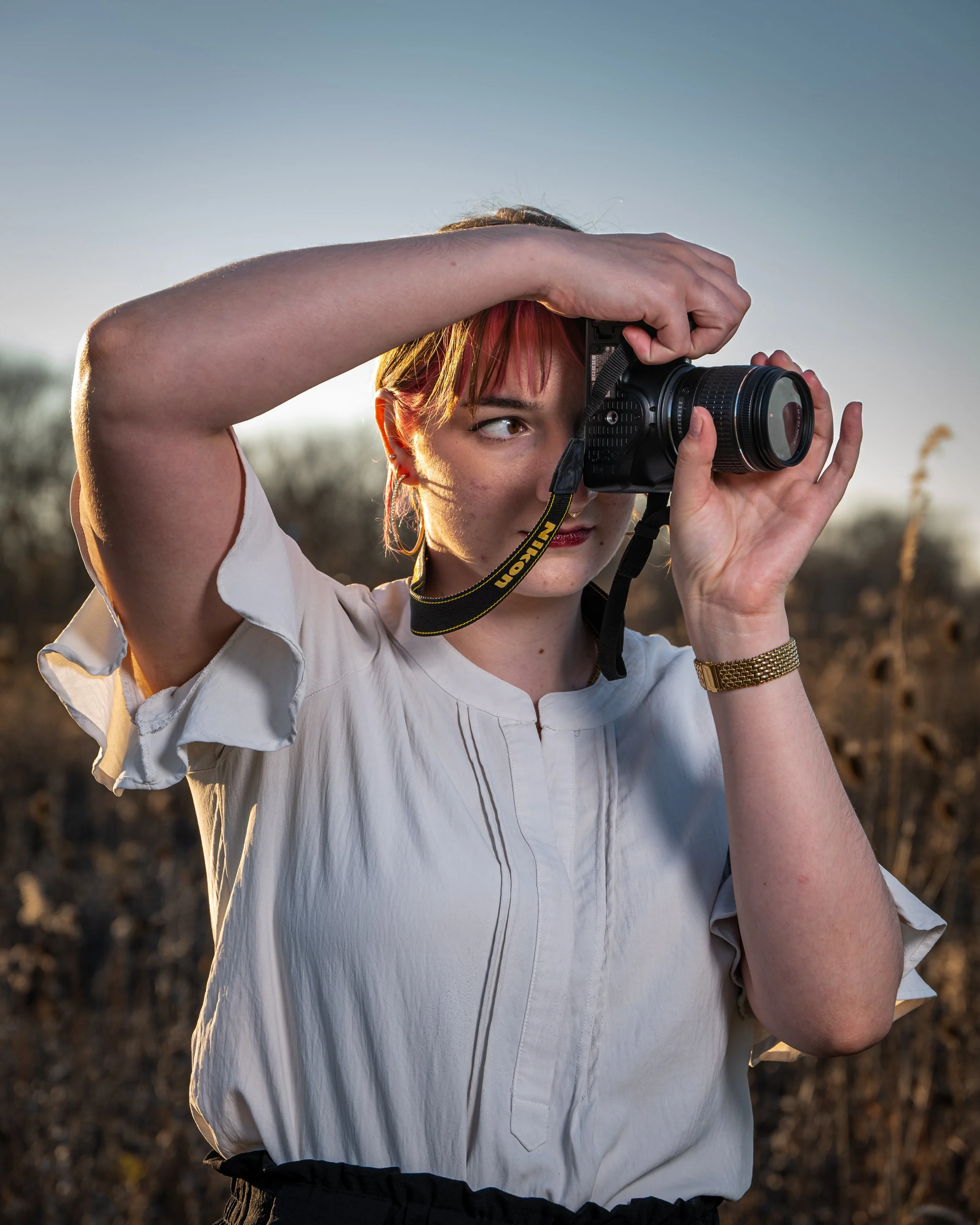 A woman with short red hair wearing a white blouse and gold watch, taking a photograph outdoors with a Nikon camera at sunset or sunrise.