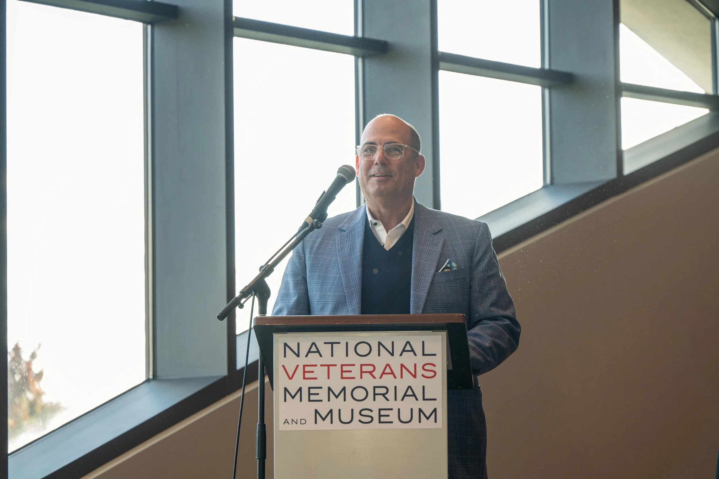 A man wearing glasses and a checked blazer stands at a podium, speaking into a microphone inside a building with large windows in the background. The podium has a sign that reads 'National Veterans Memorial and Museum'.