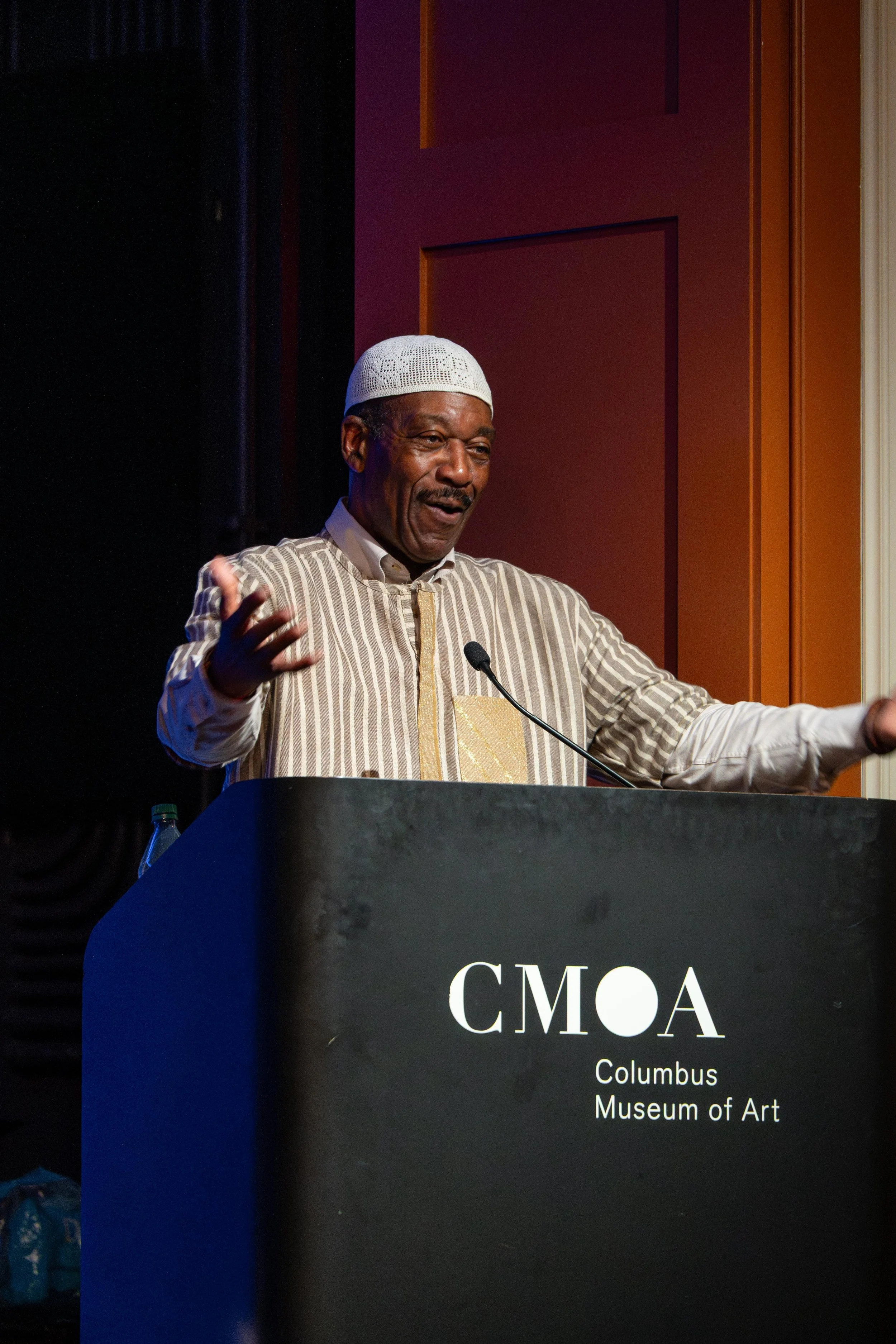 A man wearing a white kufi cap and a striped traditional outfit speaking at a podium with a microphone at the Columbus Museum of Art. The podium has the logo 'CMA' and the location 'Columbus Museum of Art' written on it.
