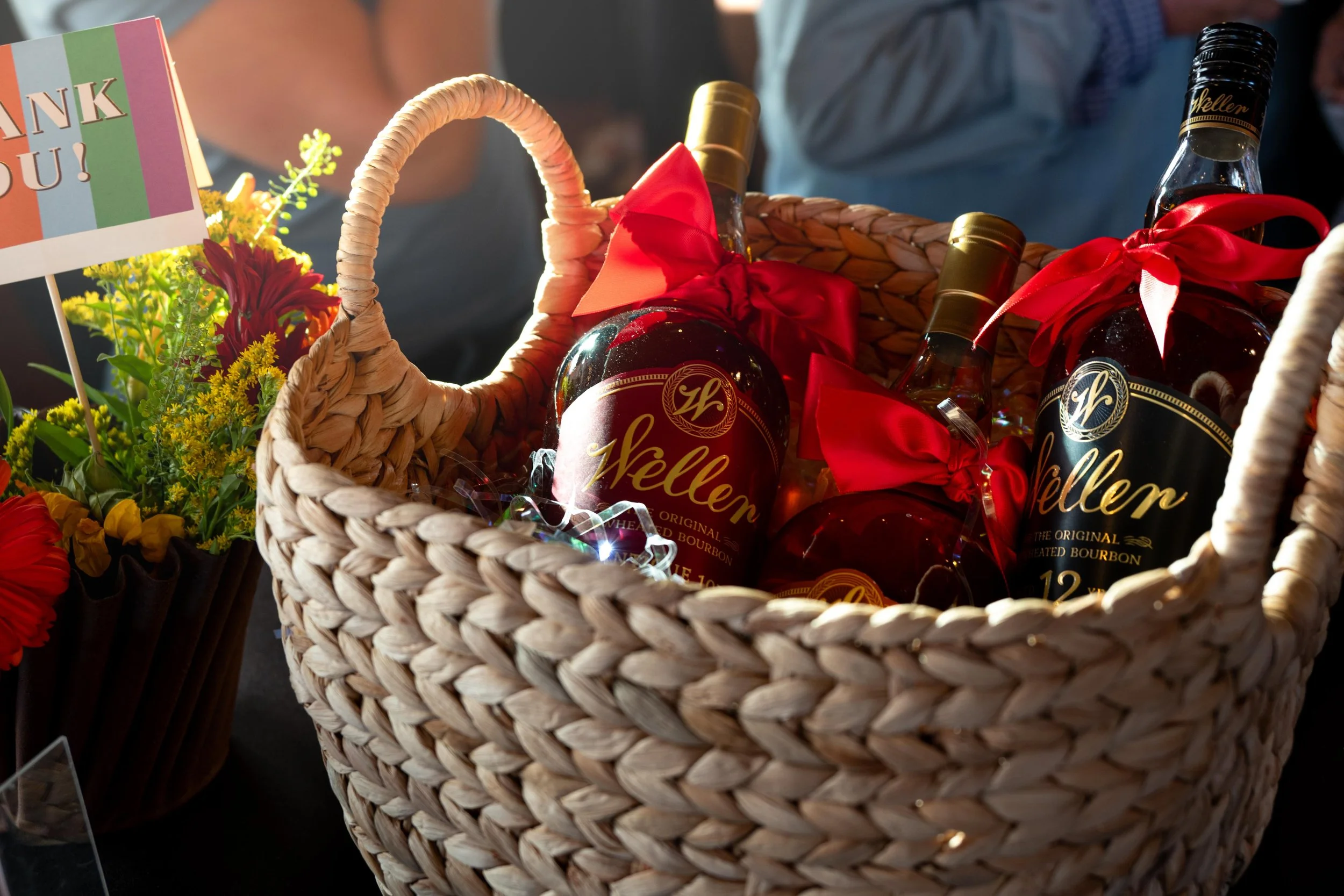 A woven basket filled with bottles of alcohol, decorations, and a 'Thank You!' card, along with flowers nearby.