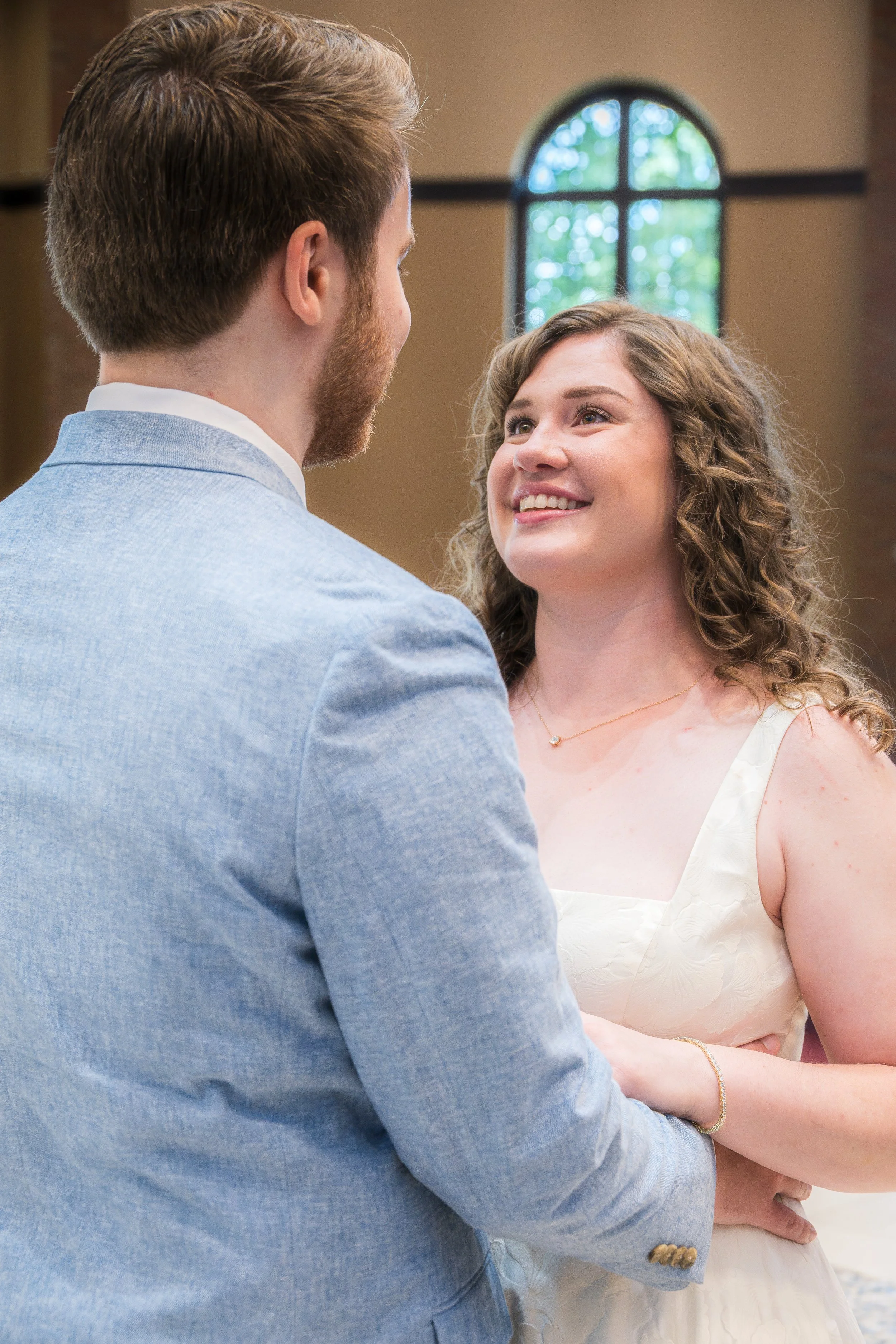 A couple holding hands and smiling at each other during their wedding ceremony inside a church with a large arched window in the background.
