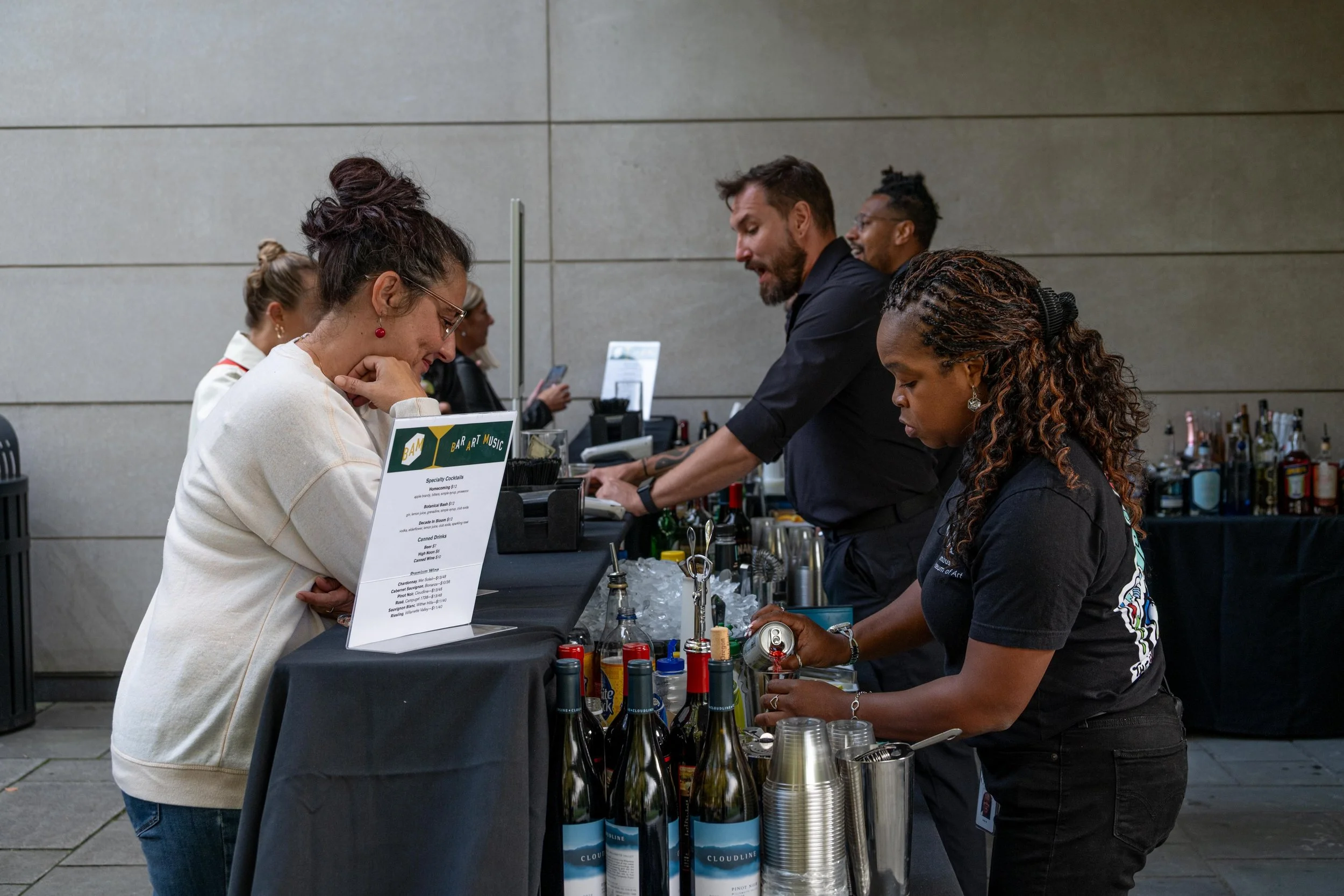 People at a bar or cocktail stand, engaging in conversation and preparing drinks, with alcohol bottles and a menu displayed on the counter.