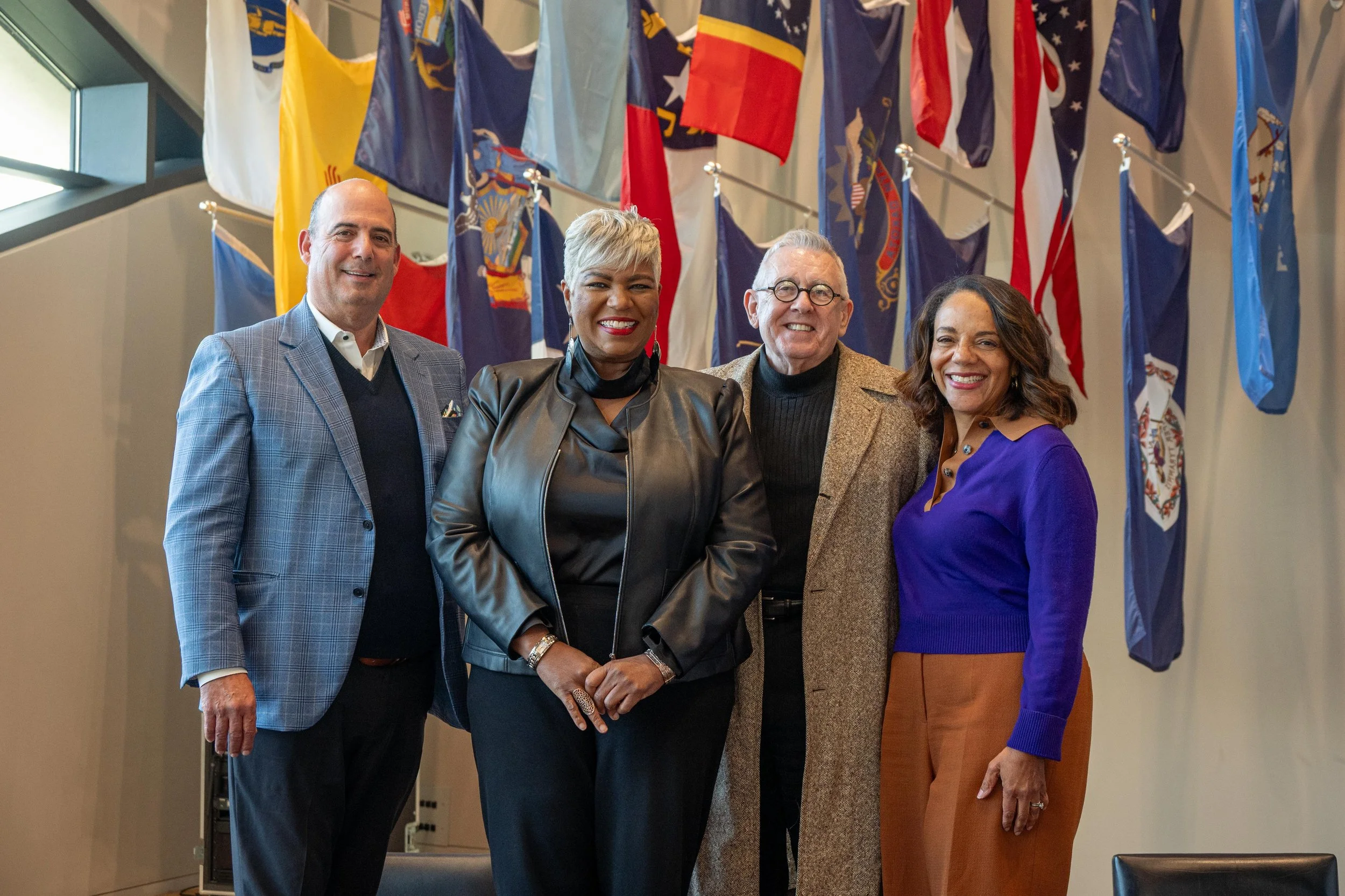 Four people standing in front of a display of various international flags, smiling for a photo. The group includes two men and two women, dressed in business attire.