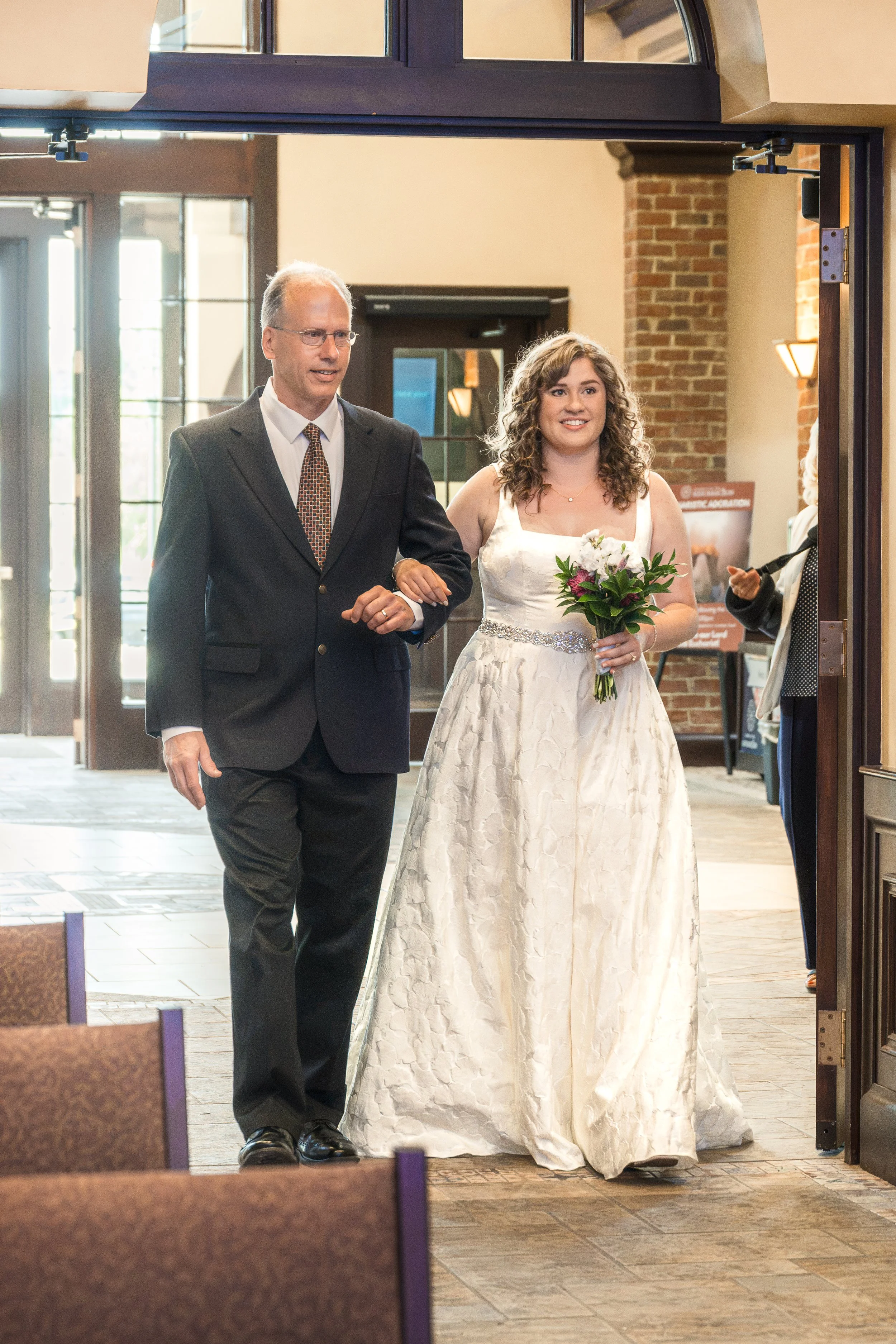 A bride in a white wedding dress holding a bouquet of flowers walking into a room, accompanied by an older man in a dark suit and glasses, with brick and glass doors in the background.