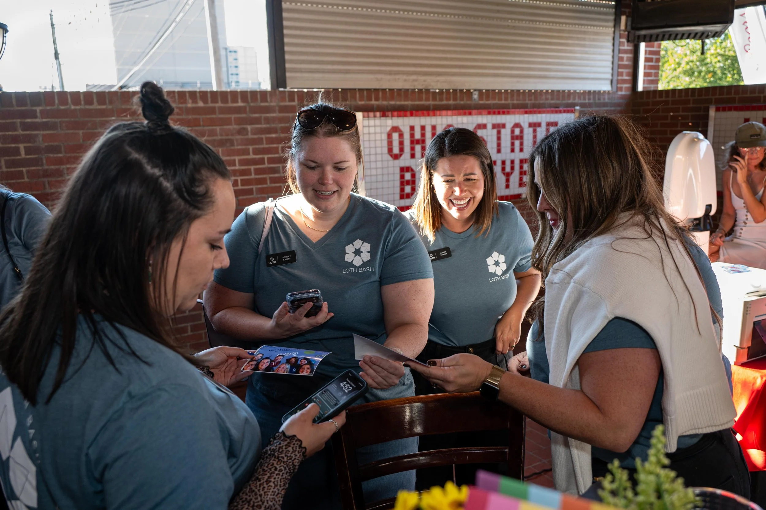 Group of women gathered around a table indoors, looking at photos and using phones, at an event with a sign in the background reading 'OHIO STATE DAY'.