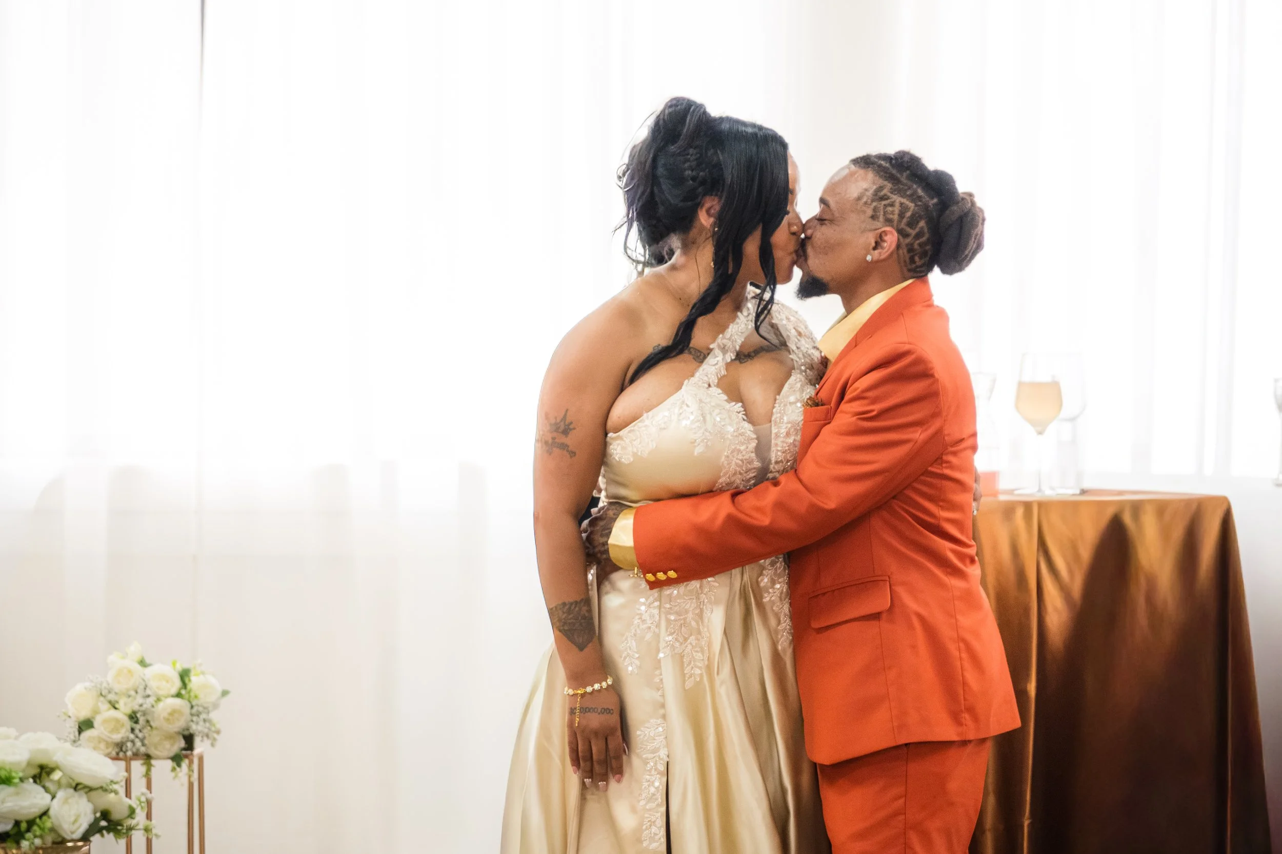 A couple kissing at their wedding ceremony, with a bride in a lace gown and a groom in an orange suit, standing in a bright indoor setting.