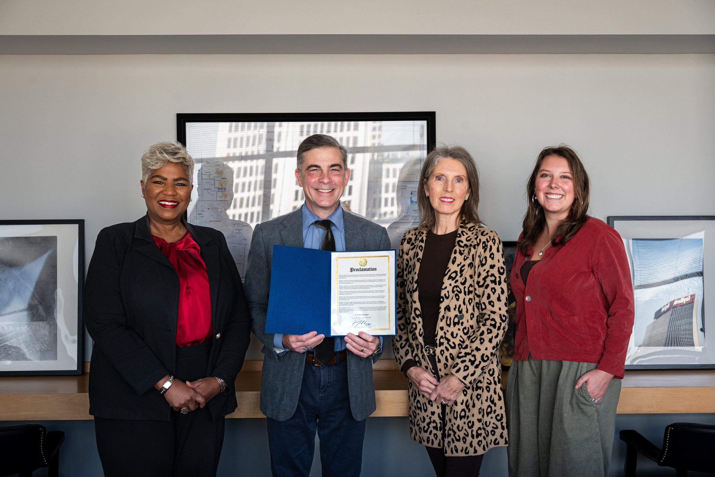 A diverse group of four adults standing indoors, with a man in the center holding a certificate. The woman on the left has short gray hair and is wearing a black blazer with a red blouse, the man in the center has gray hair, is dressed in a gray suit