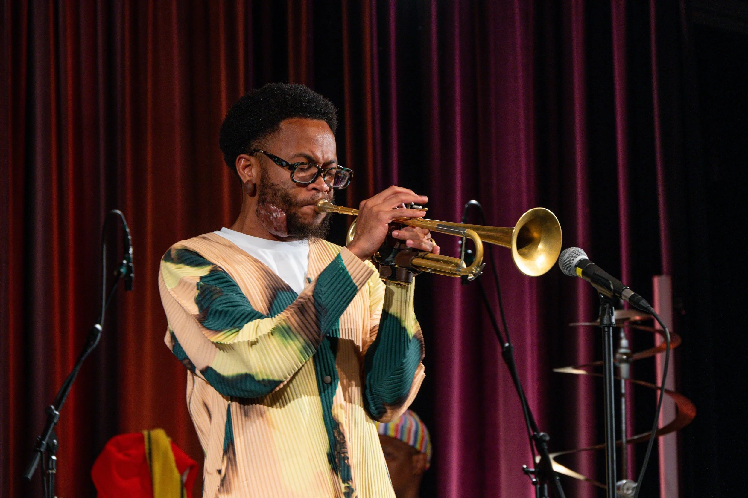 A man with glasses and a beard playing a trumpet on stage with red curtains in the background.