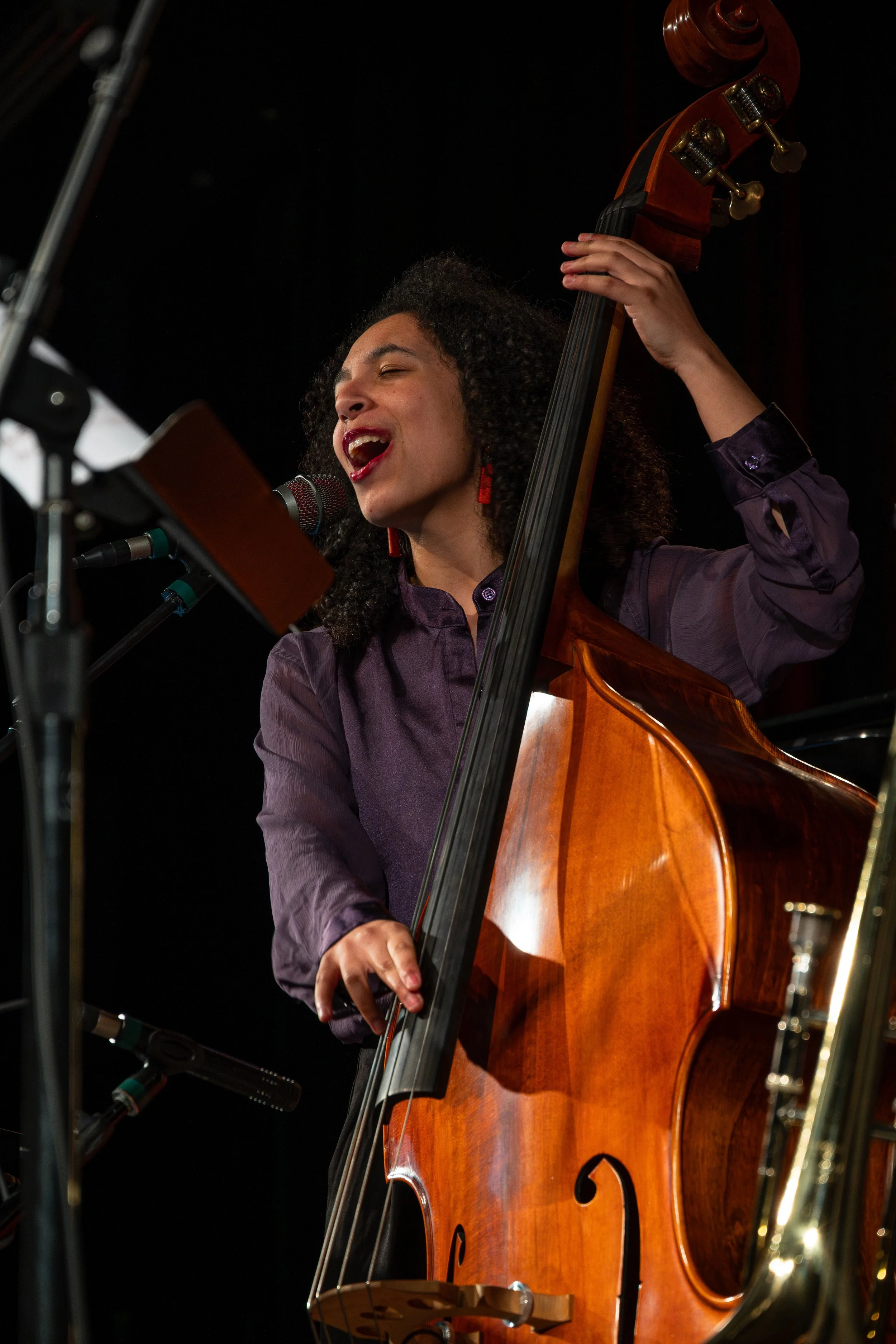 A woman singing and playing a wooden double bass during a performance, with her eyes closed and mouth open, in front of a black background.