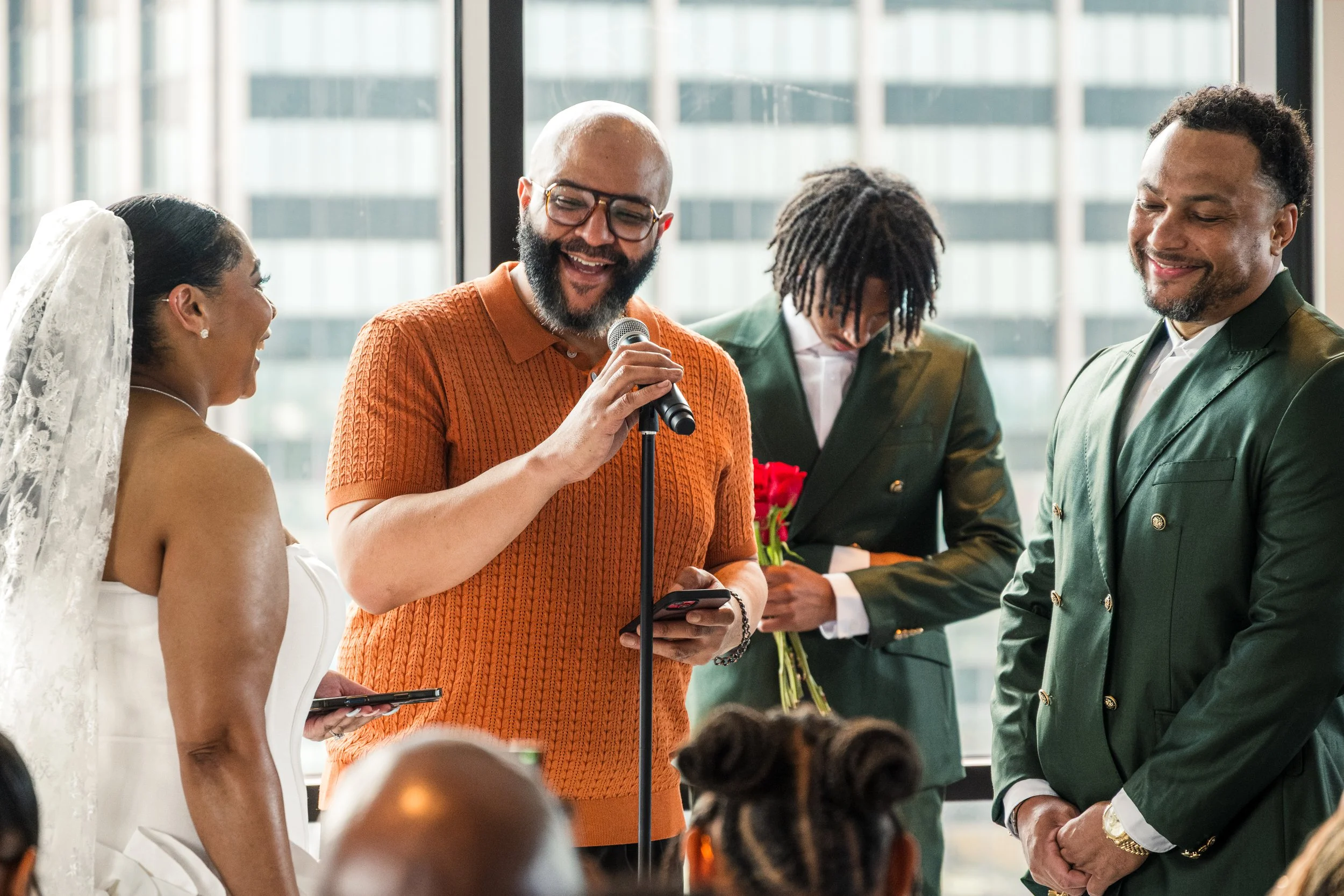 A group of diverse people at a wedding ceremony with a bride, groom, and officiant holding a microphone, standing in a bright indoor space with large windows.