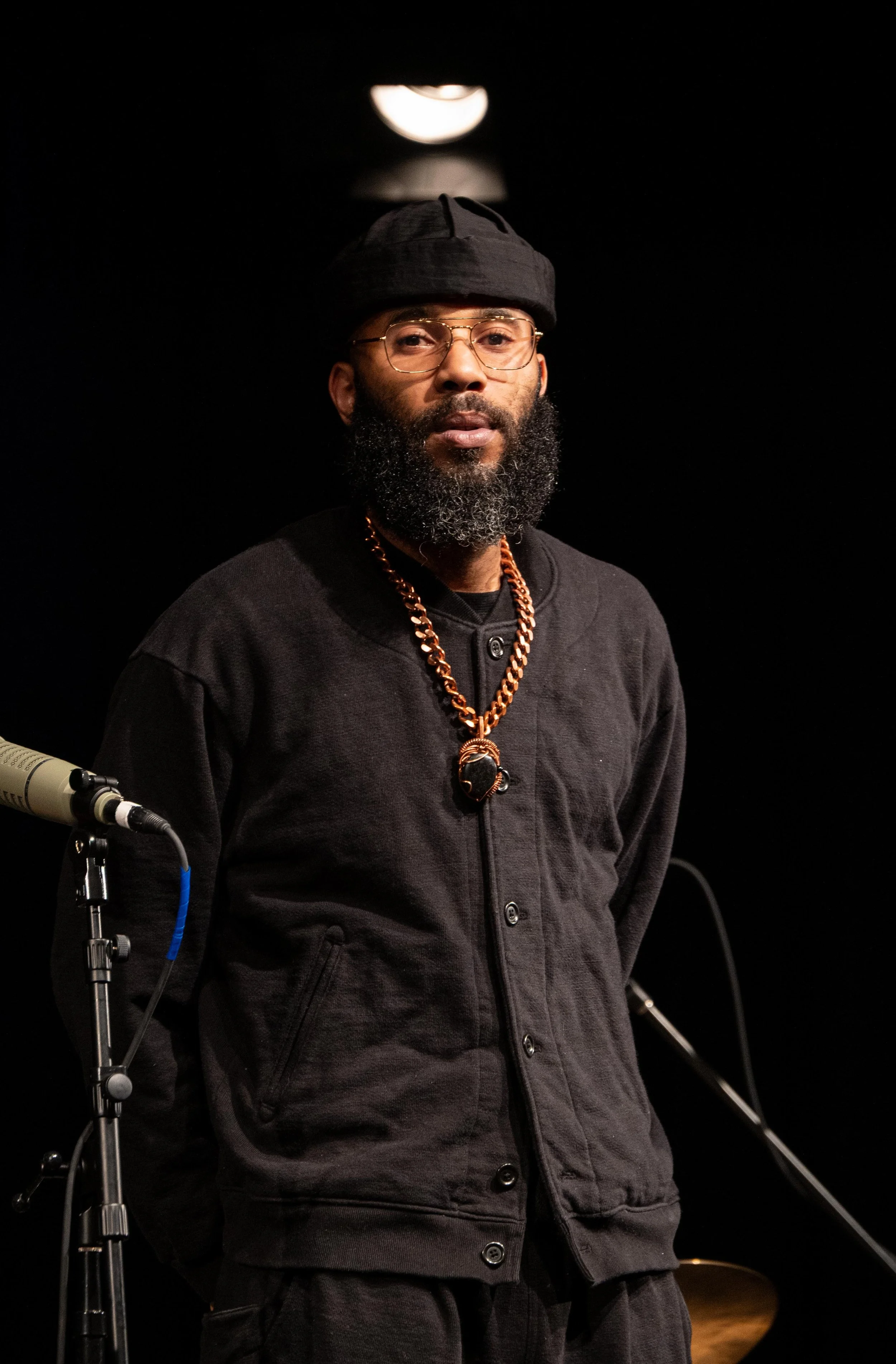A man with a beard wearing glasses, a black beanie, a black jacket, and a gold chain necklace, standing in front of a black background with a microphone on a stand and a white drum set partially visible.