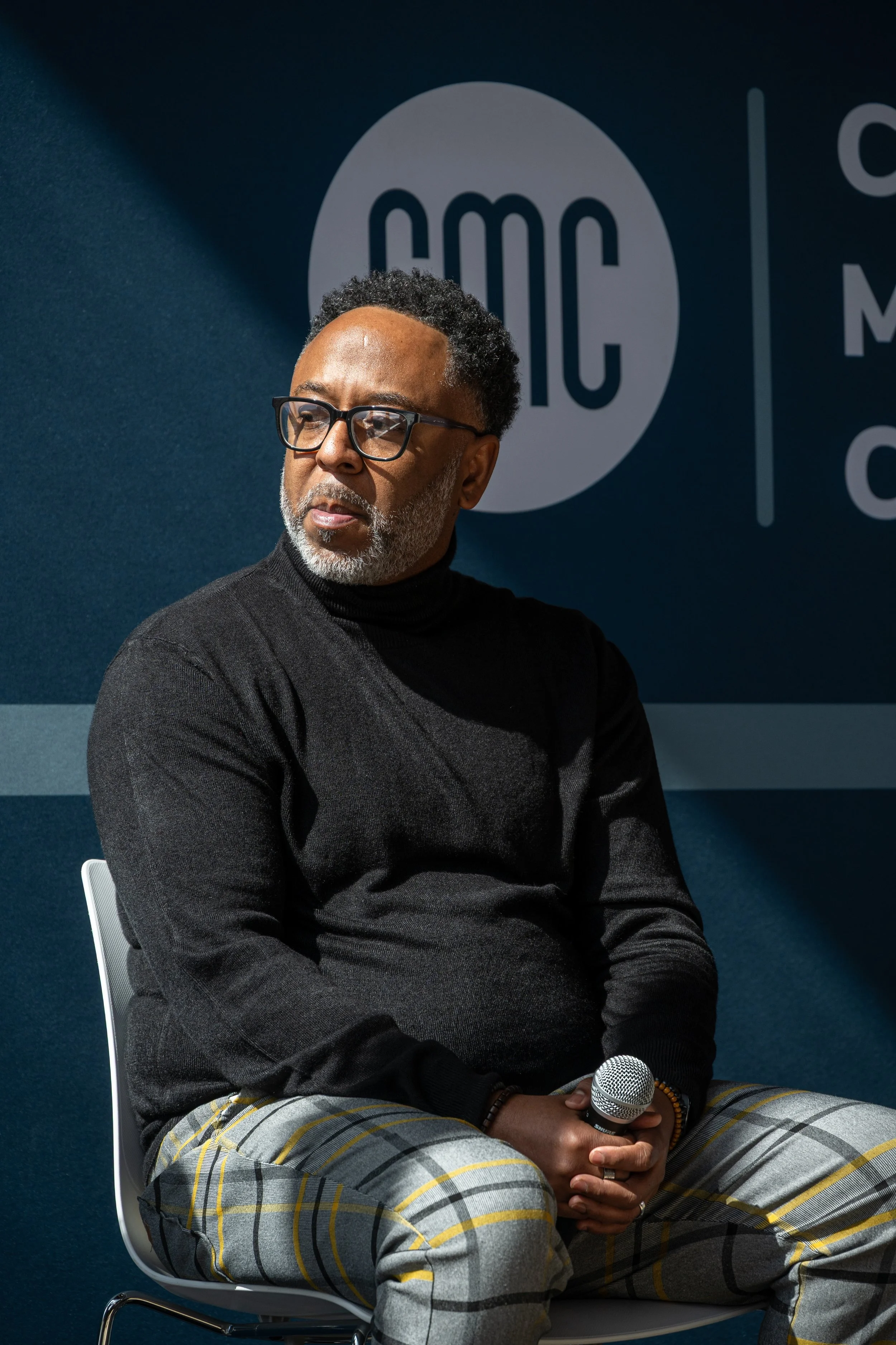 A man with glasses and a gray beard sitting in a chair holding a microphone, wearing a black turtleneck and plaid pants, in front of a backdrop with the AMC logo.