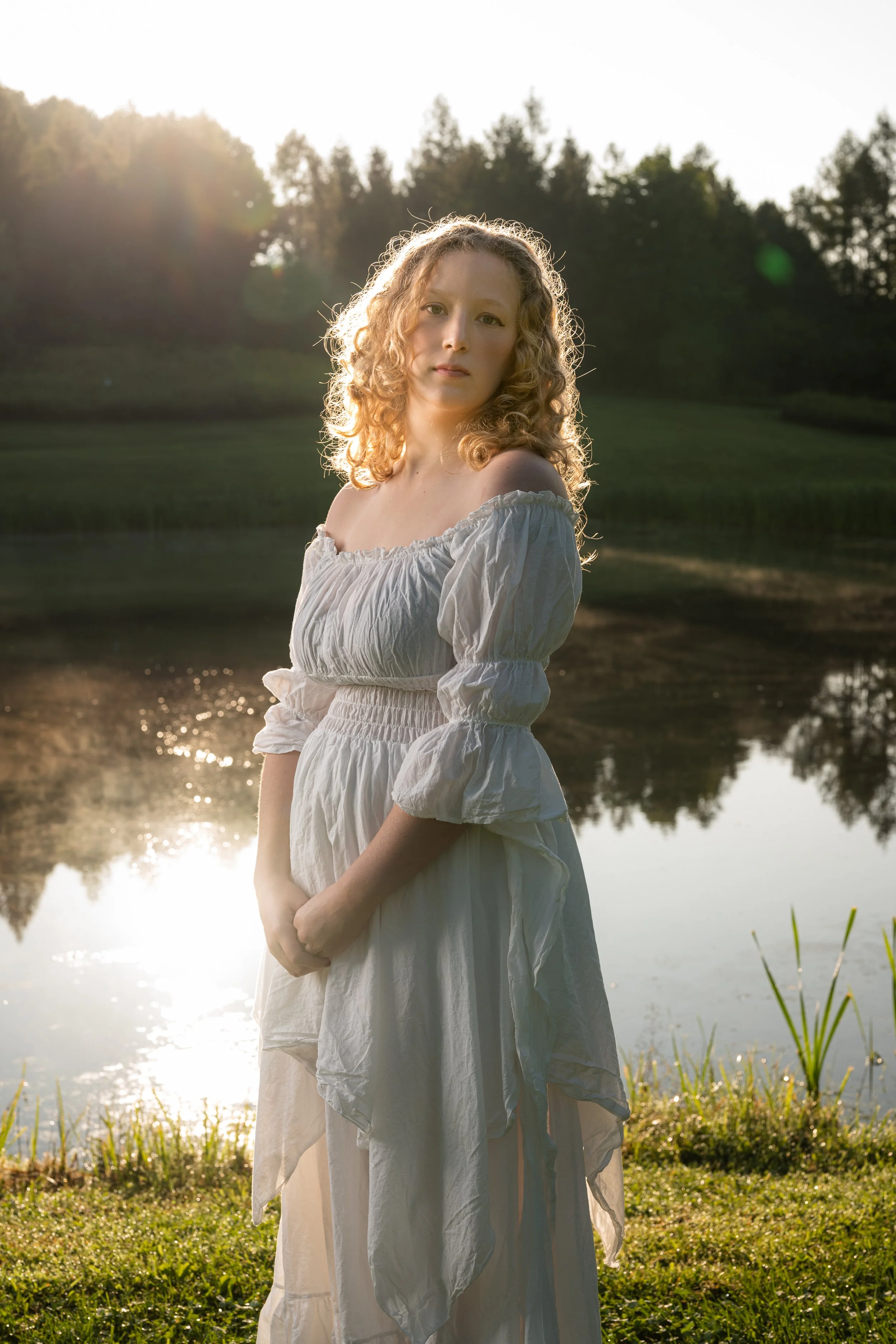 A woman with curly blonde hair standing by a lake at sunset, wearing a white off-the-shoulder dress. The background features trees and the sun shining through, creating a warm glow and reflections on the water.