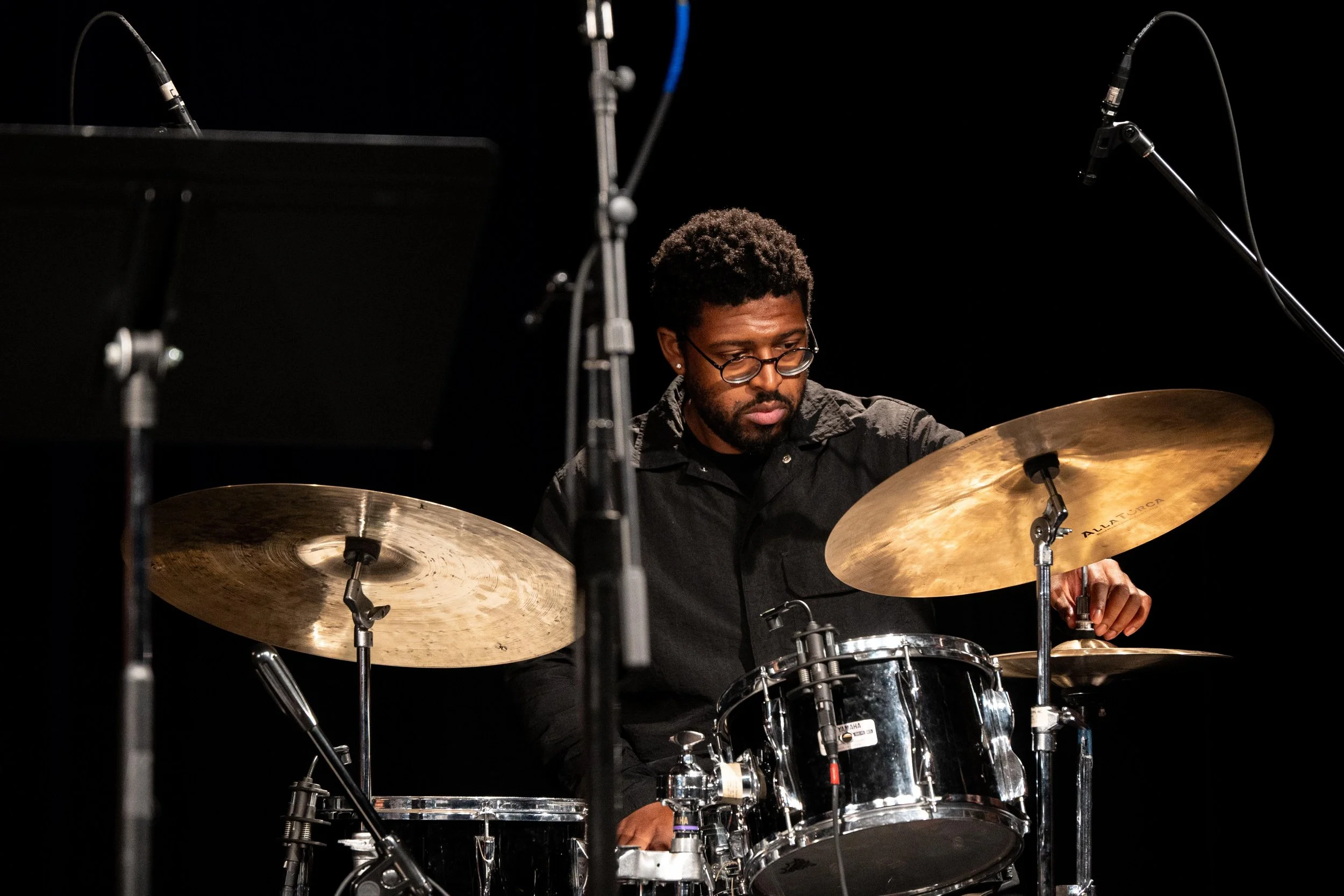 A man with glasses and curly hair playing a drum set on stage, with microphones and dark background.