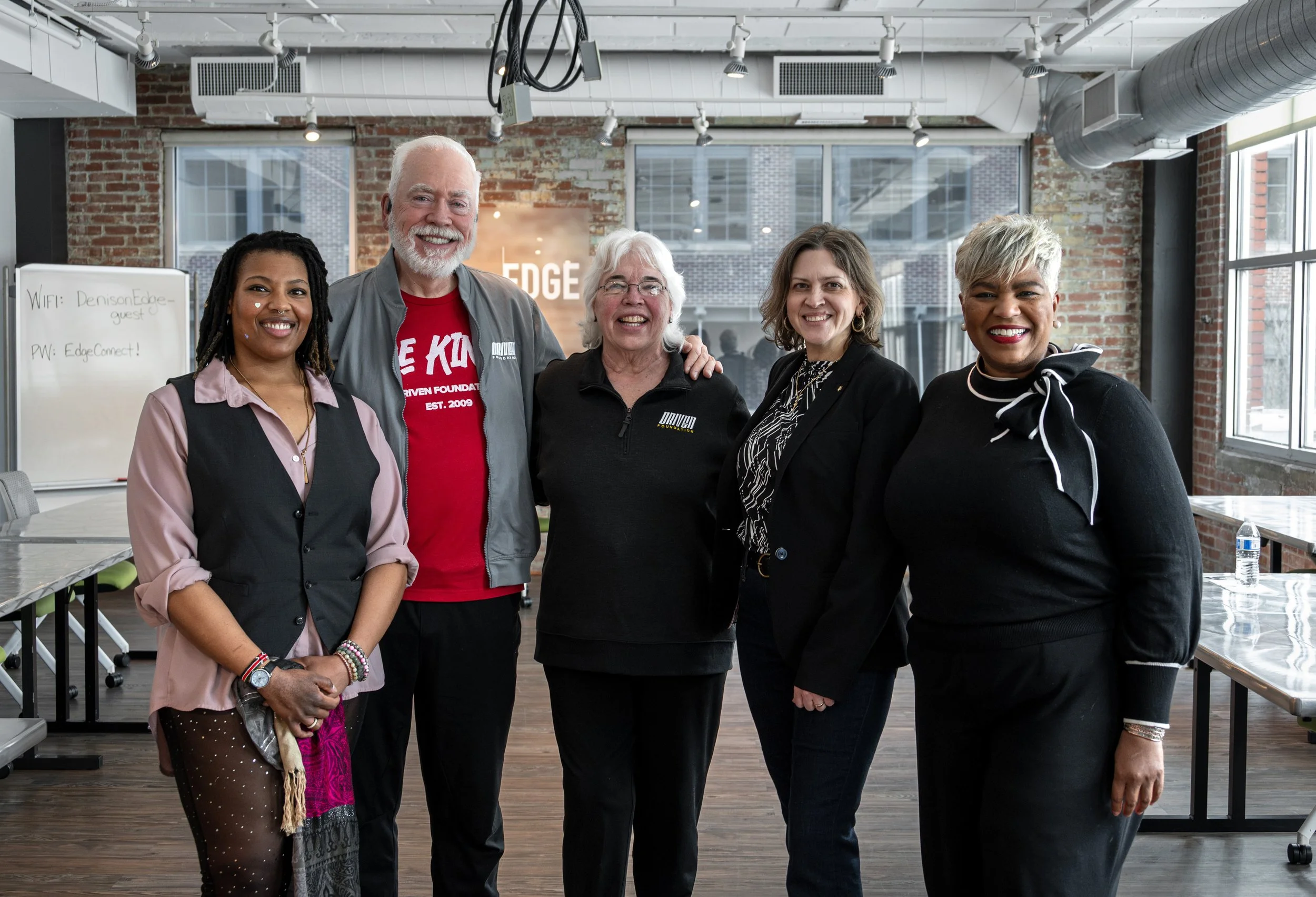 A group of five diverse smiling people standing together in a modern office space with exposed brick walls and large windows.