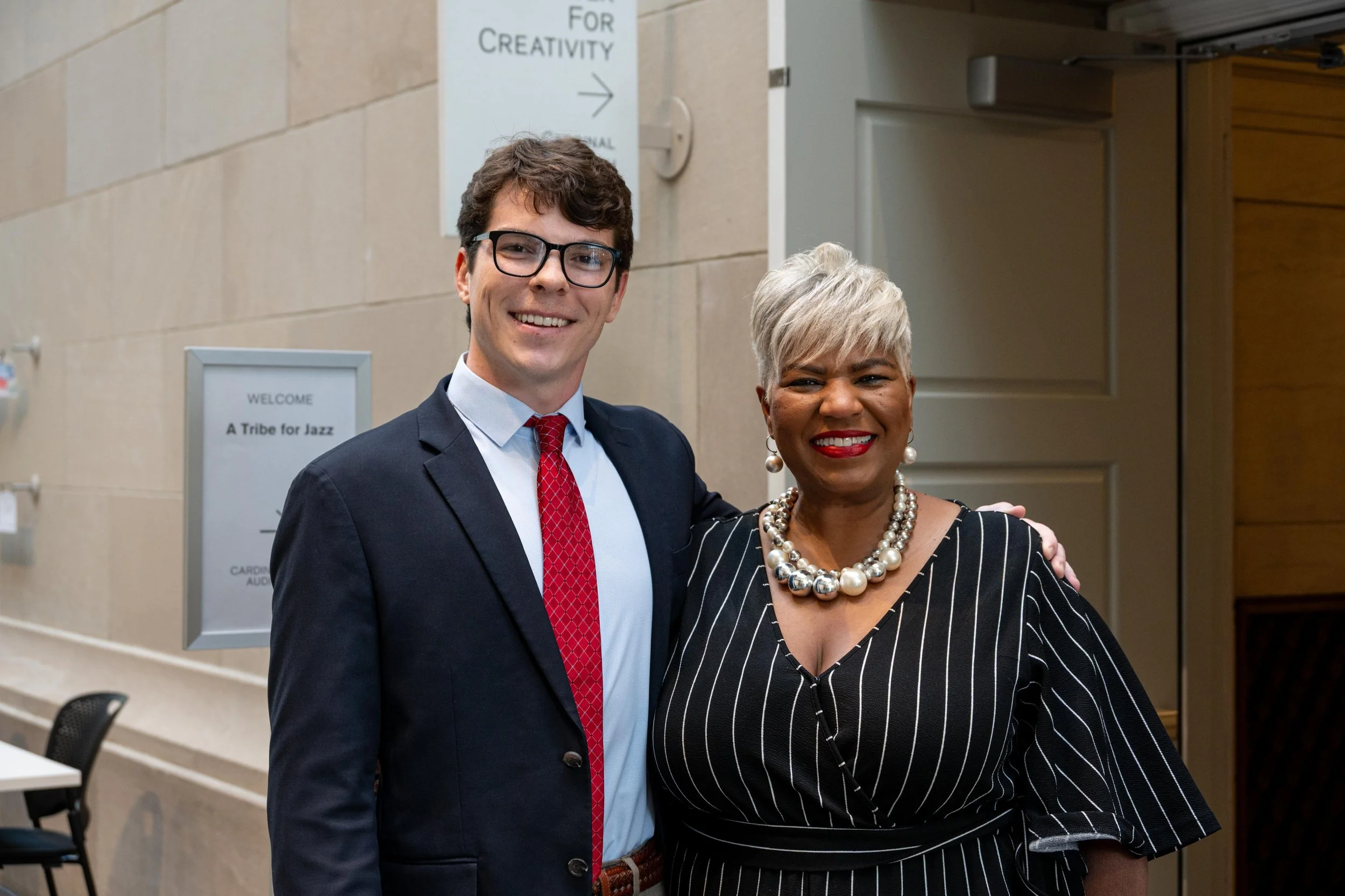A young man dressed in a dark suit, light blue shirt, and red tie stands smiling next to an older woman with short, platinum blonde hair wearing a black and white striped dress and large pearl necklace at an indoor event.