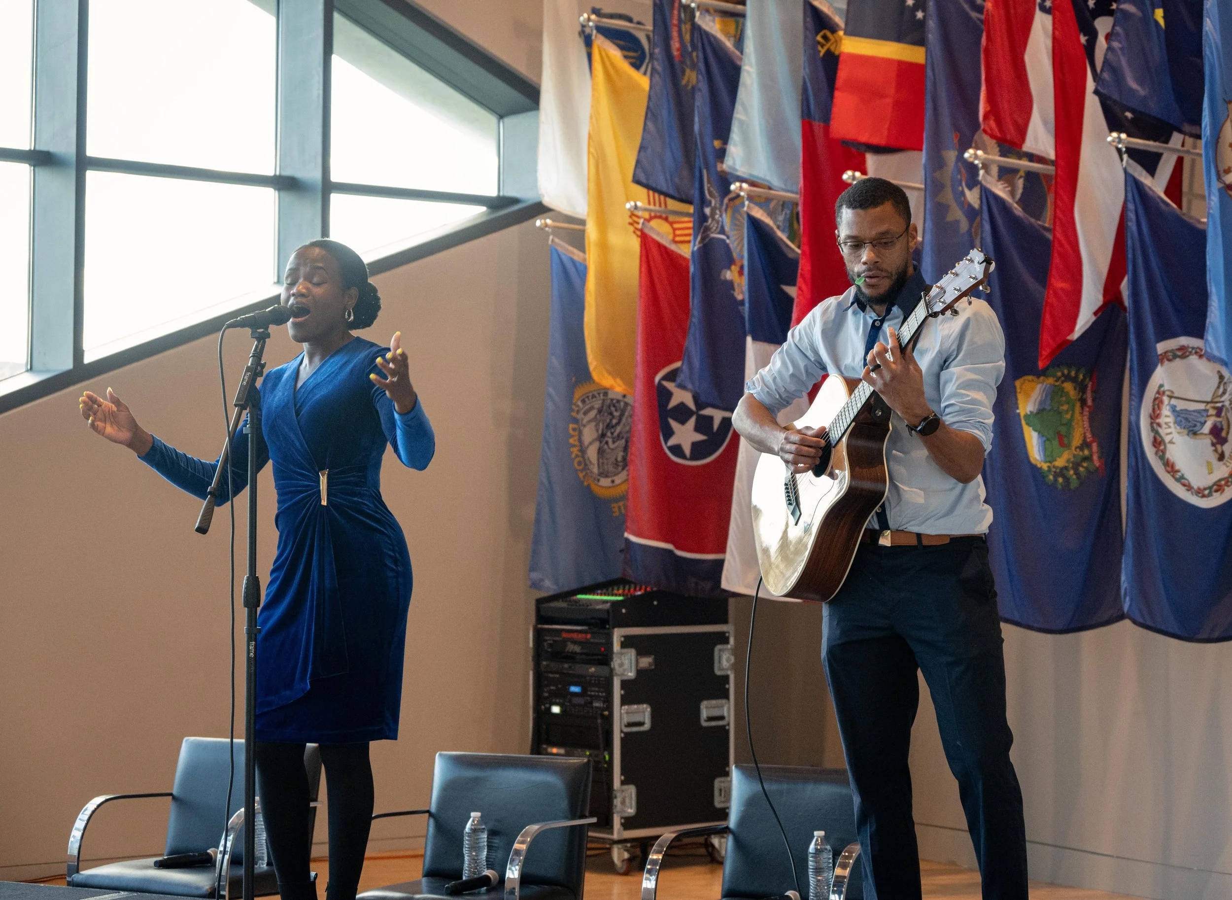 A woman singing into a microphone and a man playing an acoustic guitar during a performance, with multiple colorful flags hanging on the wall behind them.