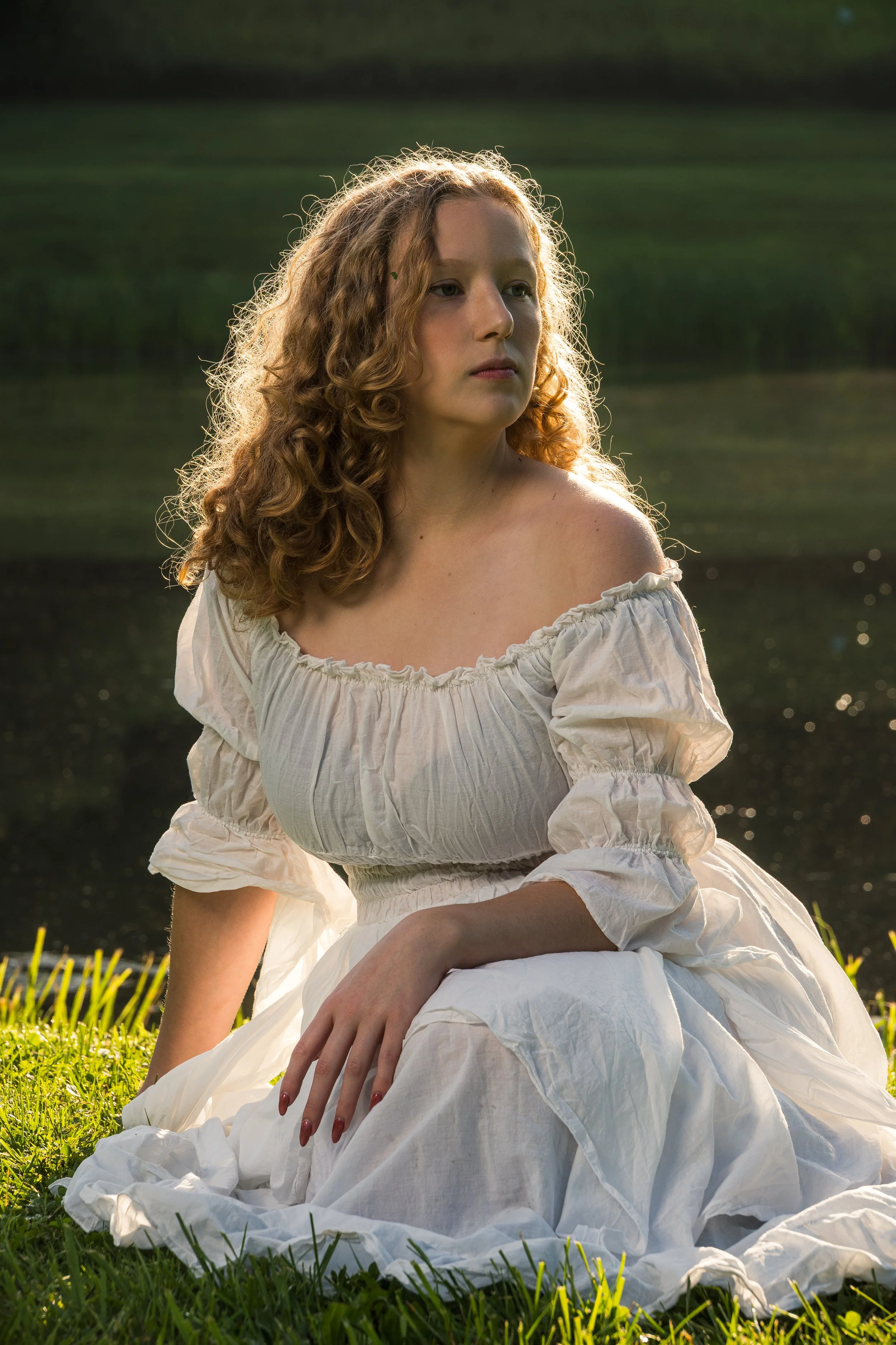 A young woman with curly hair, dressed in a white vintage-style dress with puffed sleeves, sits outdoors on grass near a body of water during golden hour, with soft sunlight illuminating her face and hair.