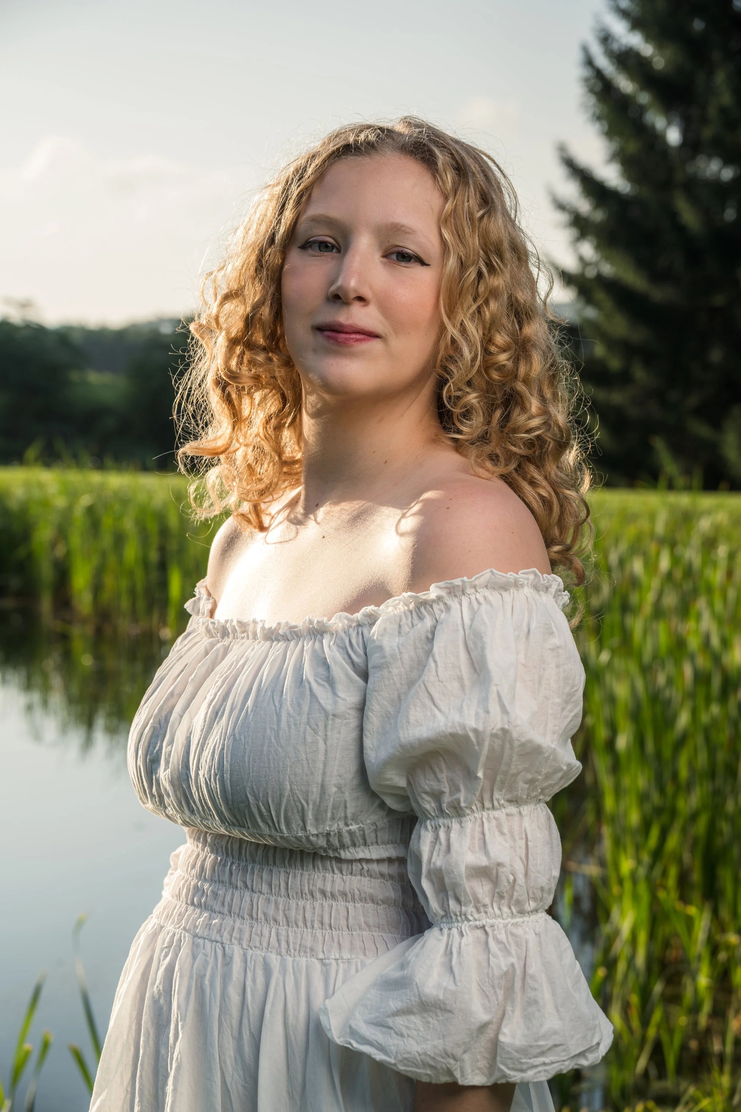 A young woman with curly blonde hair standing outdoors near a body of water, wearing an off-the-shoulder white dress, with trees and greenery in the background.