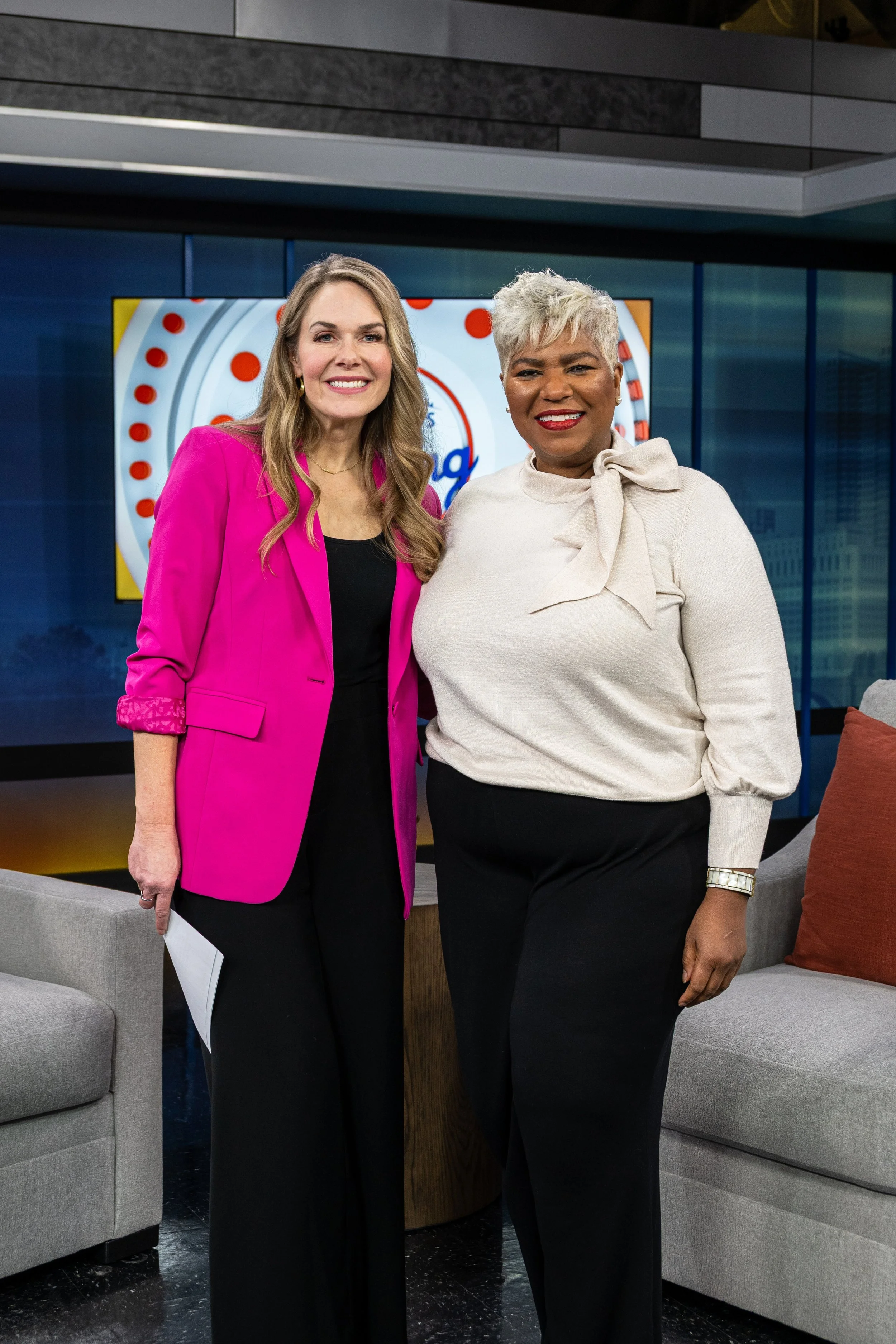 Two women standing together and smiling during a television interview or talk show. They are dressed professionally, with one woman in a bright pink blazer and the other in a cream blouse with a bow. The background shows a large screen and modern stu