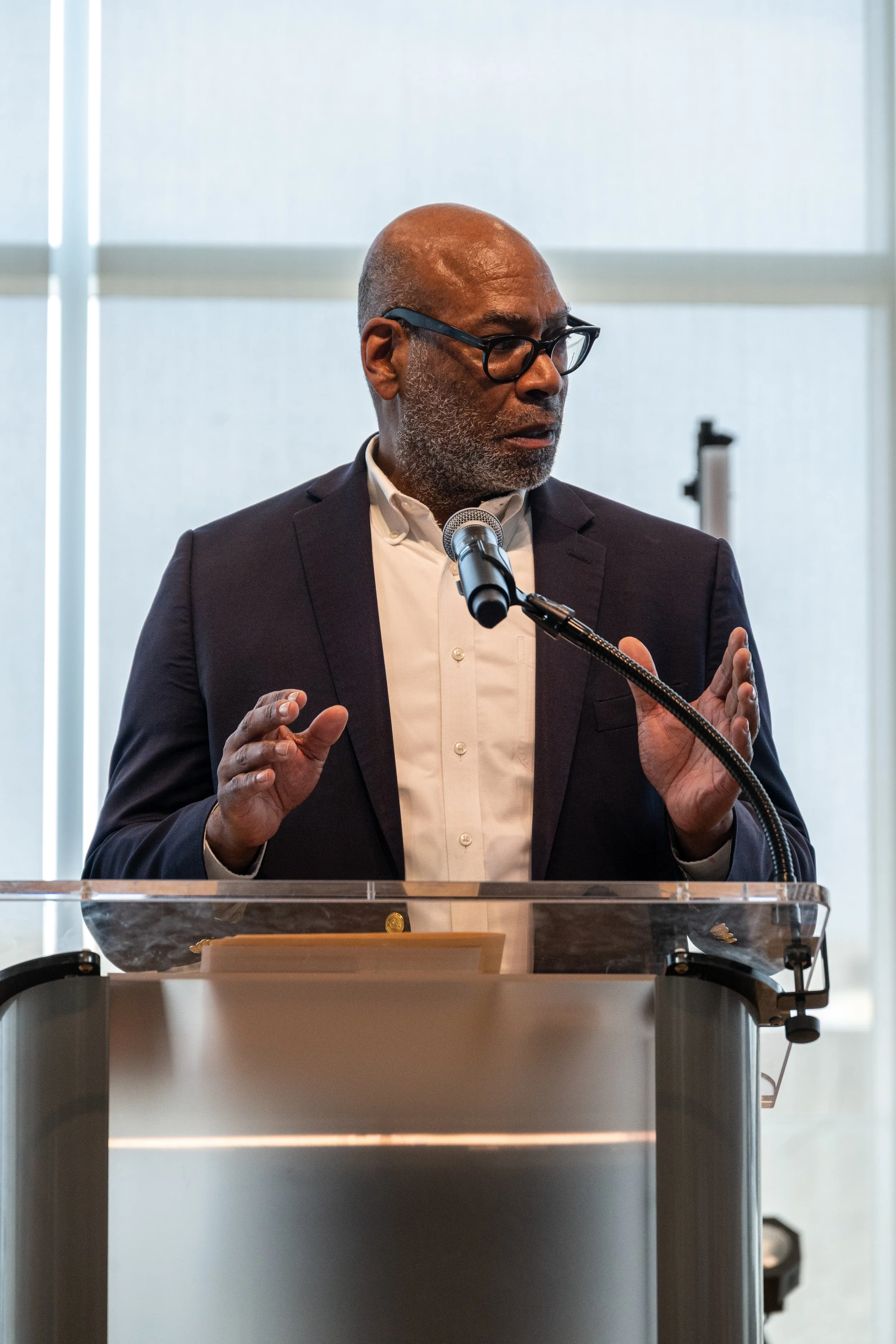 A man with dark skin, bald head, glasses, and gray beard, wearing a dark suit and white shirt, speaking from behind a clear podium with a microphone.