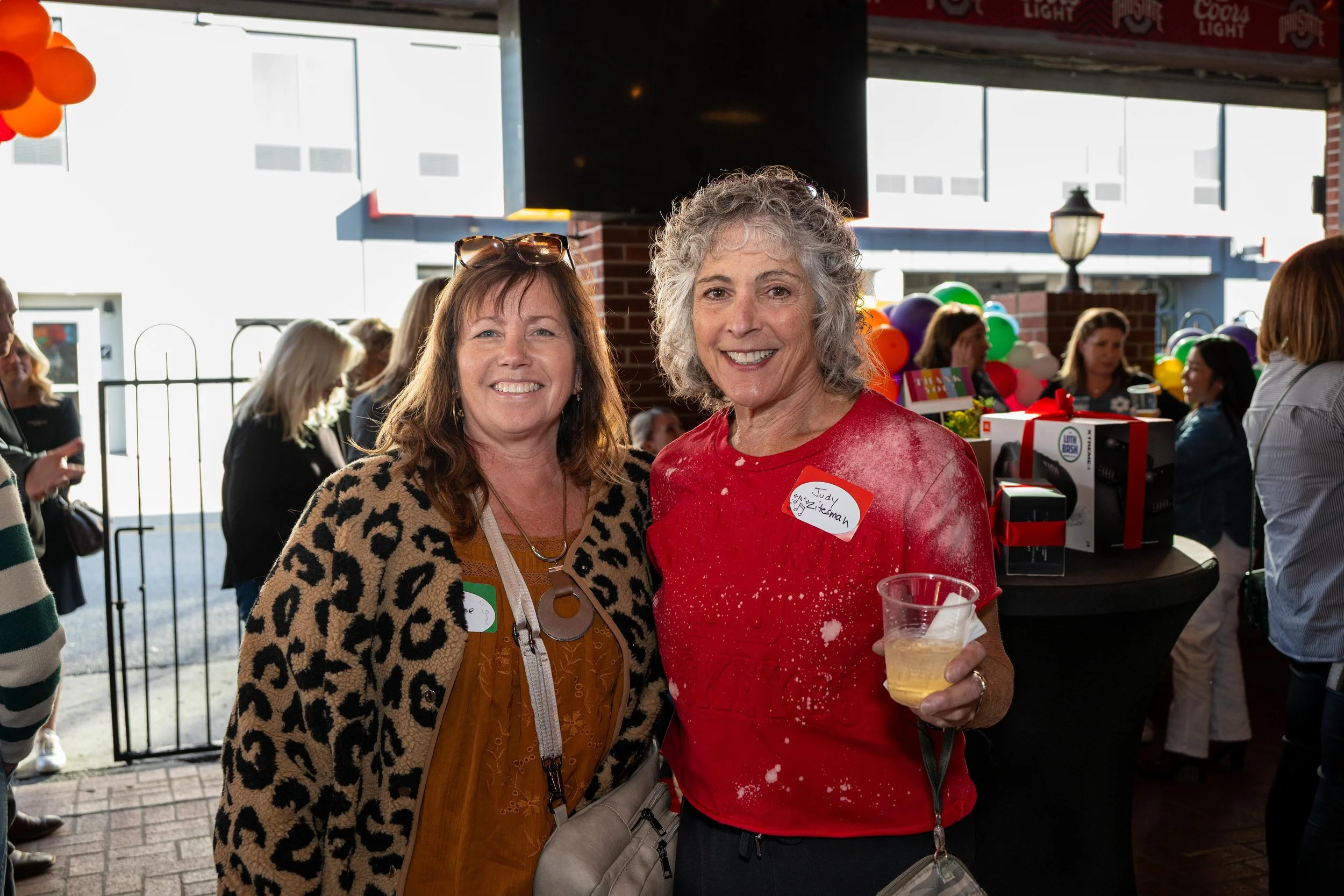 Two smiling women at a social gathering, one wearing a leopard print jacket and sunglasses on her head, the other in a red shirt with a name tag, holding a drink, with balloons and other people in the background.