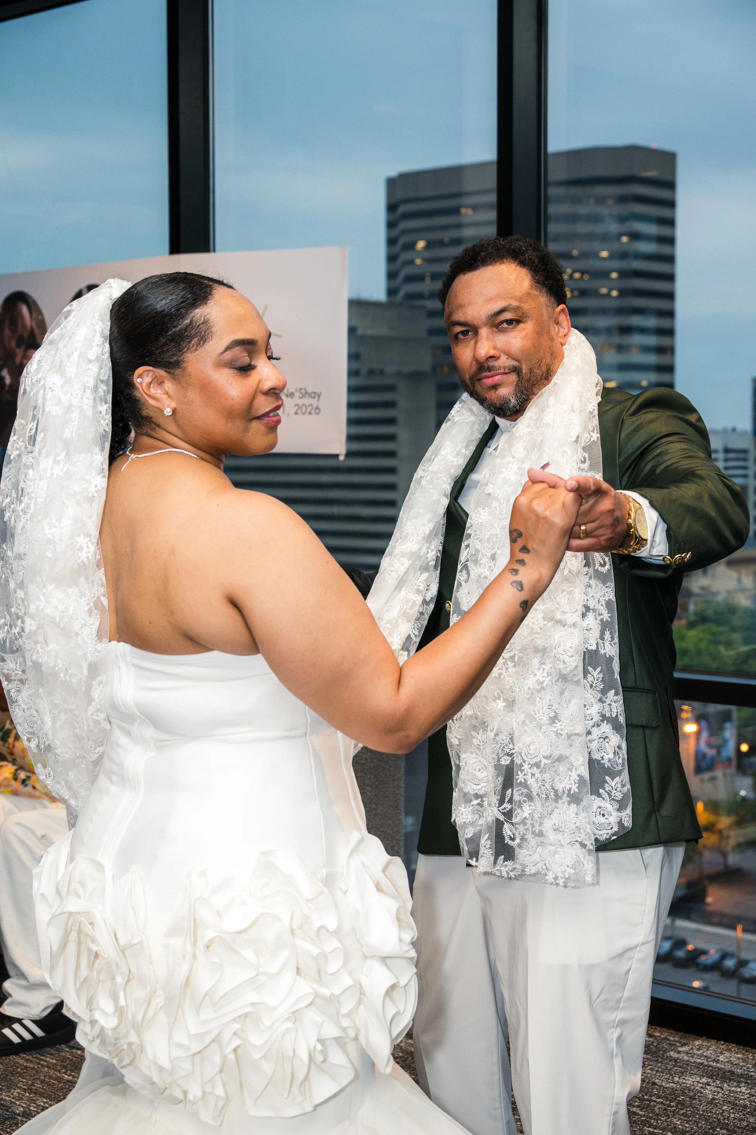 A bride and groom dance at their wedding reception in a high-rise building, with city skyscrapers visible through large windows in the background.