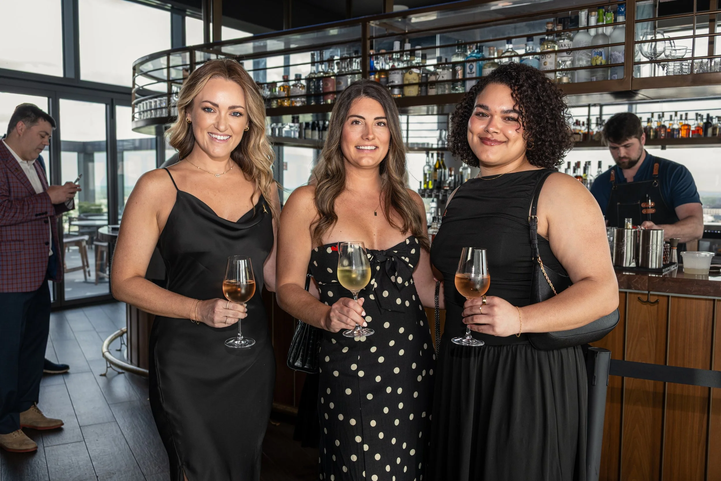 Three women standing in a bar holding wine glasses with white wine, smiling at the camera, with a bar and bartender in the background.