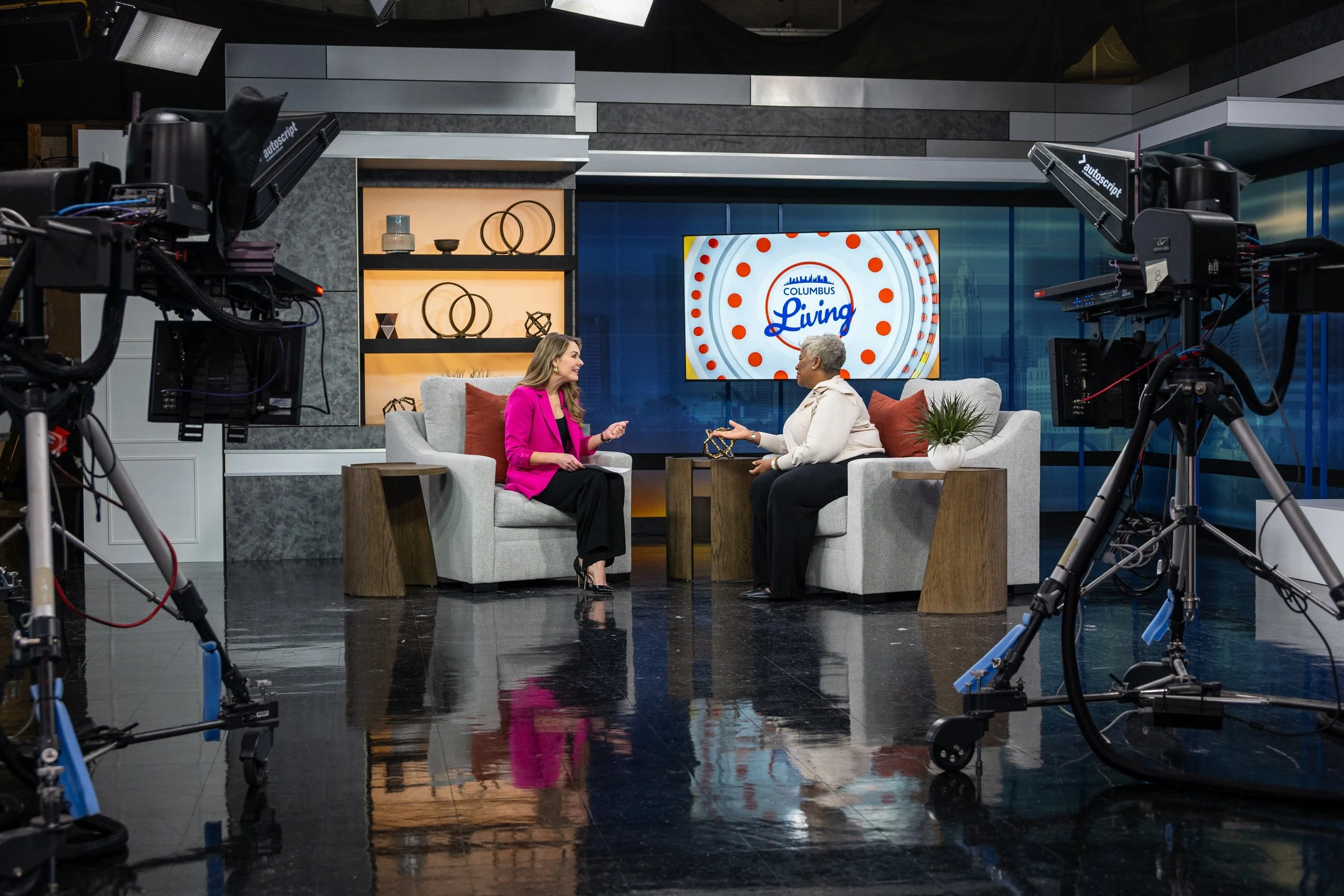 Two women sitting and talking on a television set with cameras pointed at them, a large screen behind them displays a logo that says 'Columbus Living', and decorative shelves with various objects are in the background.
