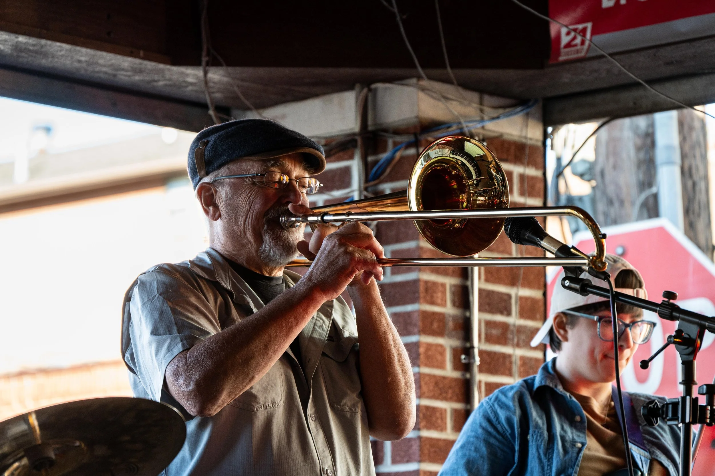 Older man playing the trombone, wearing glasses, a dark cap, and a beige shirt, with brick wall and young person with glasses and hat in the background.