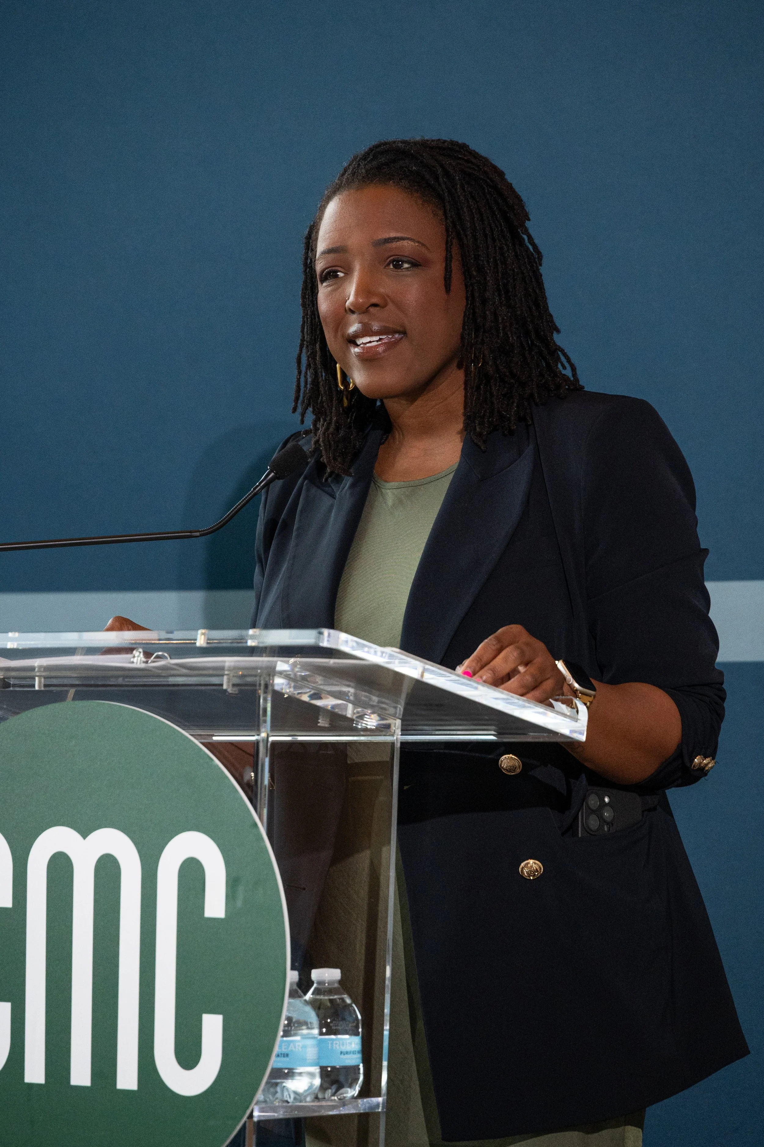 A woman with dark skin and shoulder-length dreadlocks is speaking at a podium. She is wearing a black blazer with gold buttons, a green top, and gold hoop earrings. There is a microphone attached to the podium.