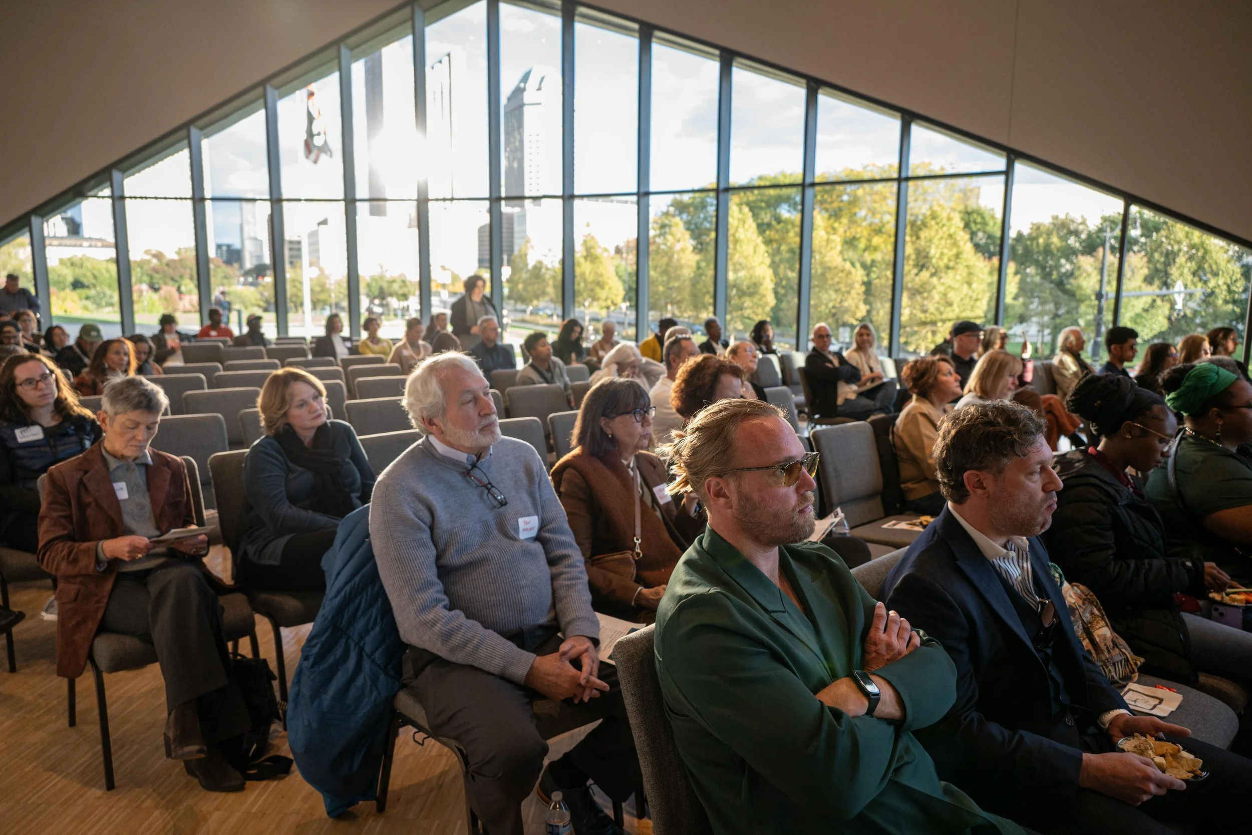 A group of people attending a conference or seminar in a modern room with large glass windows showing trees outside. The audience is seated, listening attentively, with some taking notes or holding papers.