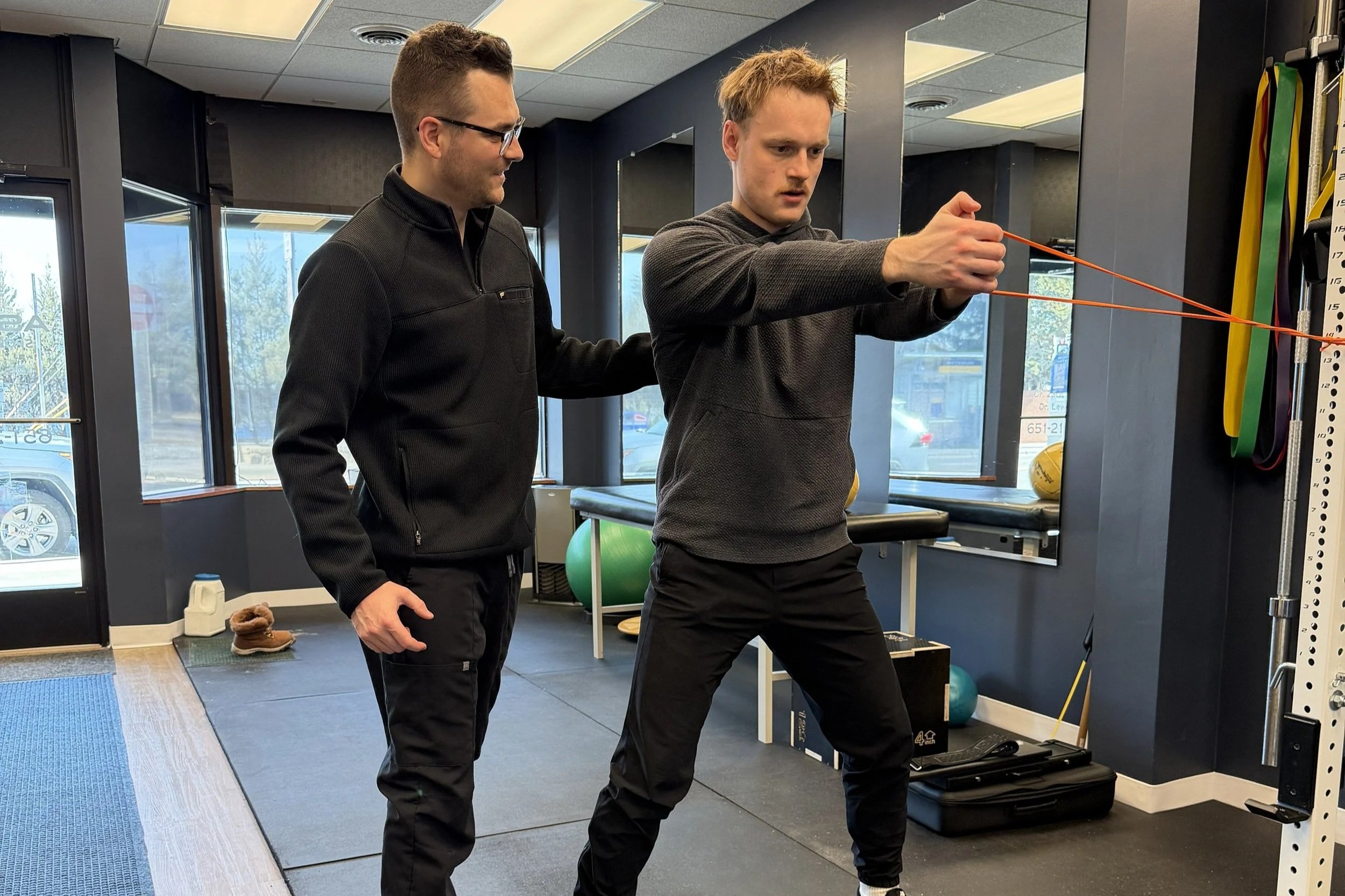 Two individuals are holding a red resistance band, with one person pulling on each end, indicating a physical therapy or exercise session.