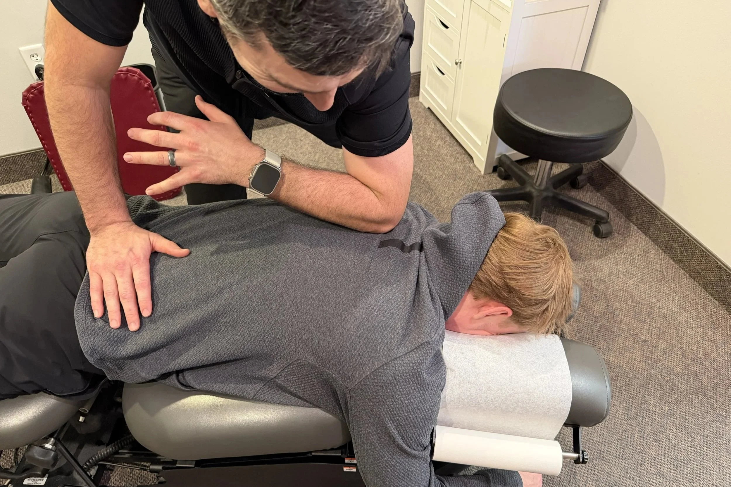 A chiropractic treatment room with a burgundy adjustable treatment table, a beige and cream granite counter with white cabinets, a model of a human spine, and a beach scene painting on the wall.