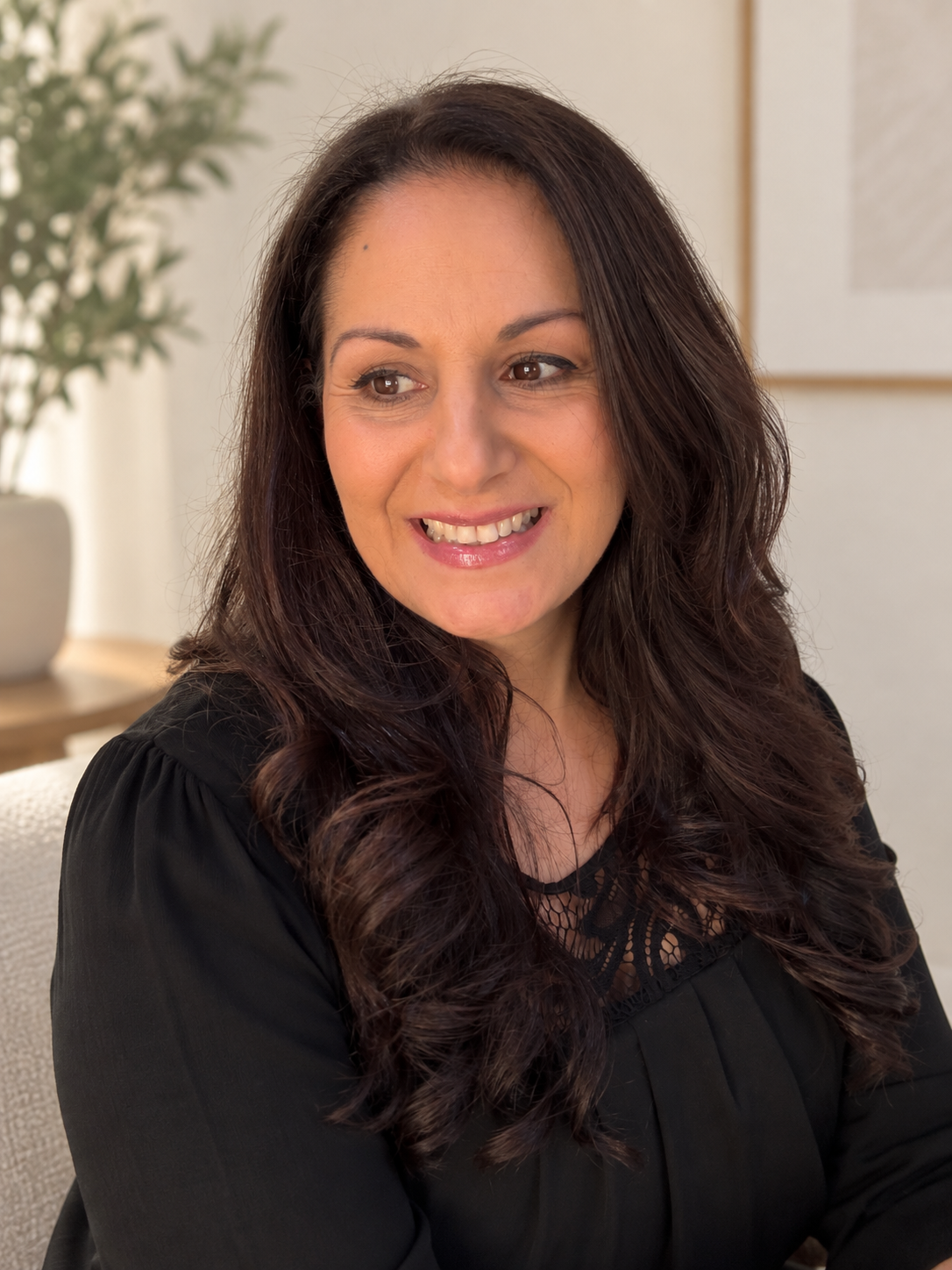 A woman with long dark brown hair smiling, wearing a black top, sitting indoors with a houseplant and framed artwork in the background.