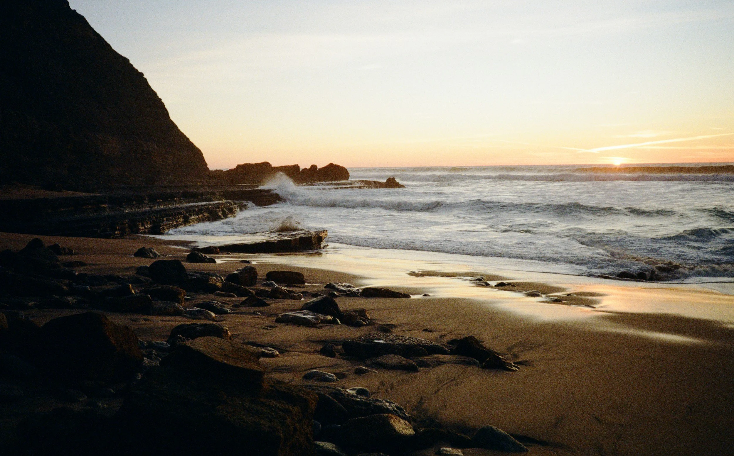 Sunset over the ocean with waves crashing onto a rocky shoreline and a large cliff on the left side.