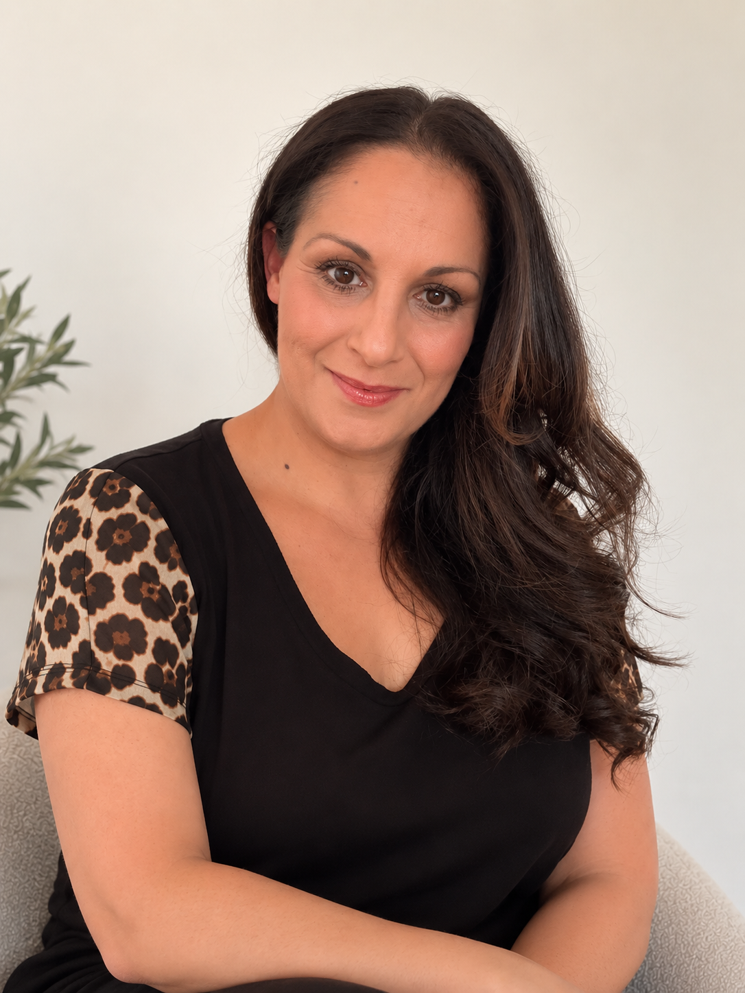 A woman with long dark hair and brown eyes, wearing a black top with leopard print sleeves, sitting on a light-colored couch near a green potted plant, smiling at the camera.