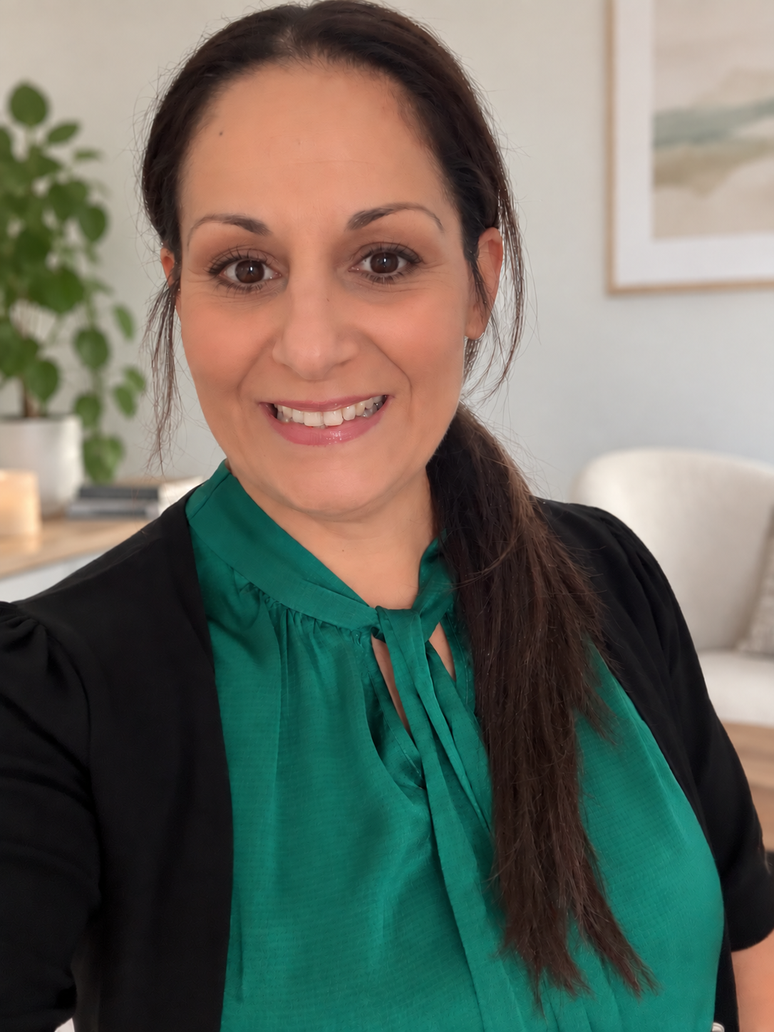 A woman with brown hair and brown eyes, smiling, wearing a green blouse with a bow at the collar, and a black jacket, in a bright room with a houseplant and framed artwork in the background.