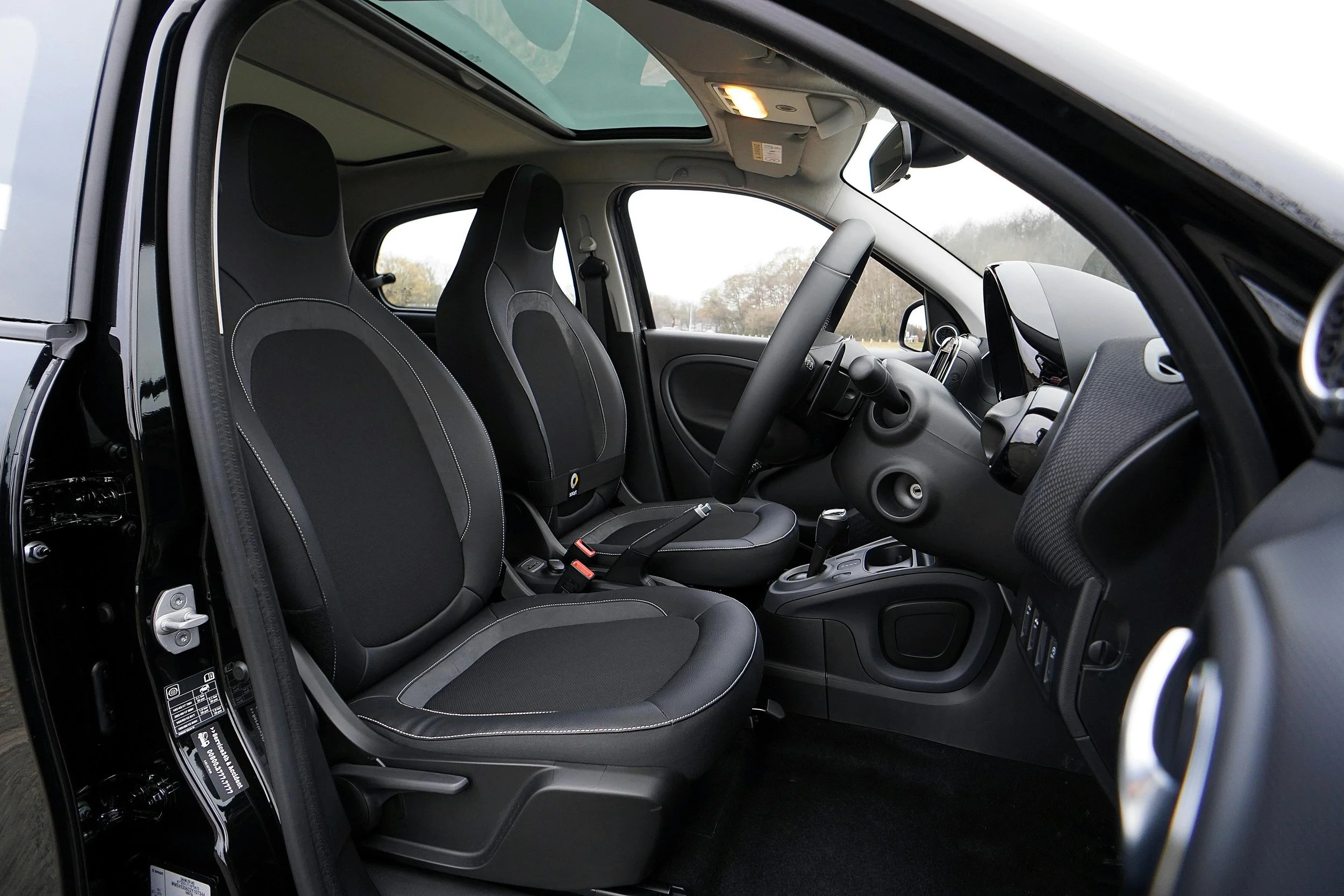 Interior of a small car showing two front seats, the dashboard, and steering wheel with a large panoramic sunroof overhead.