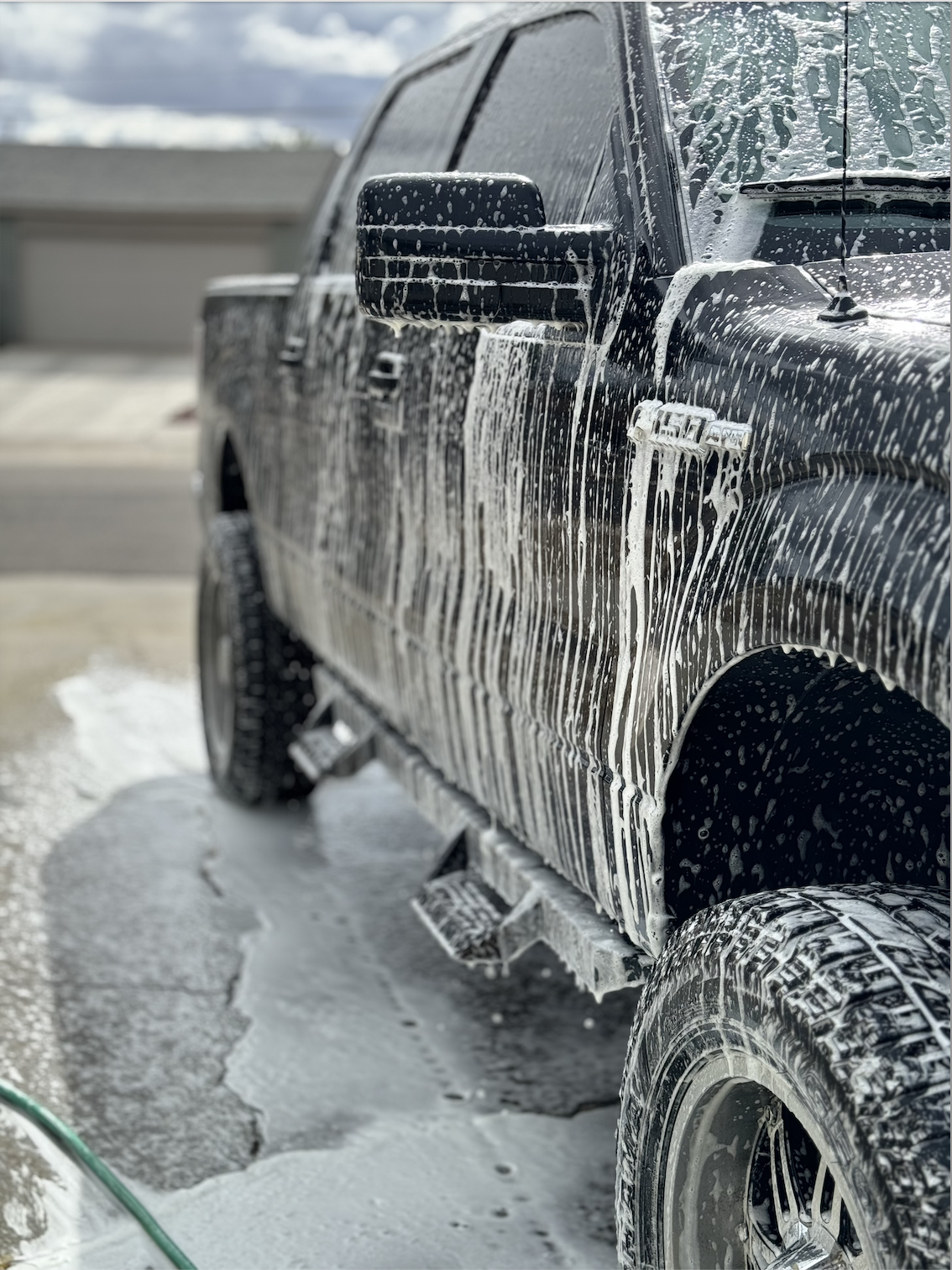 Black pickup truck being washed with soap and water, soap suds on the vehicle's surface, outside on a driveway.