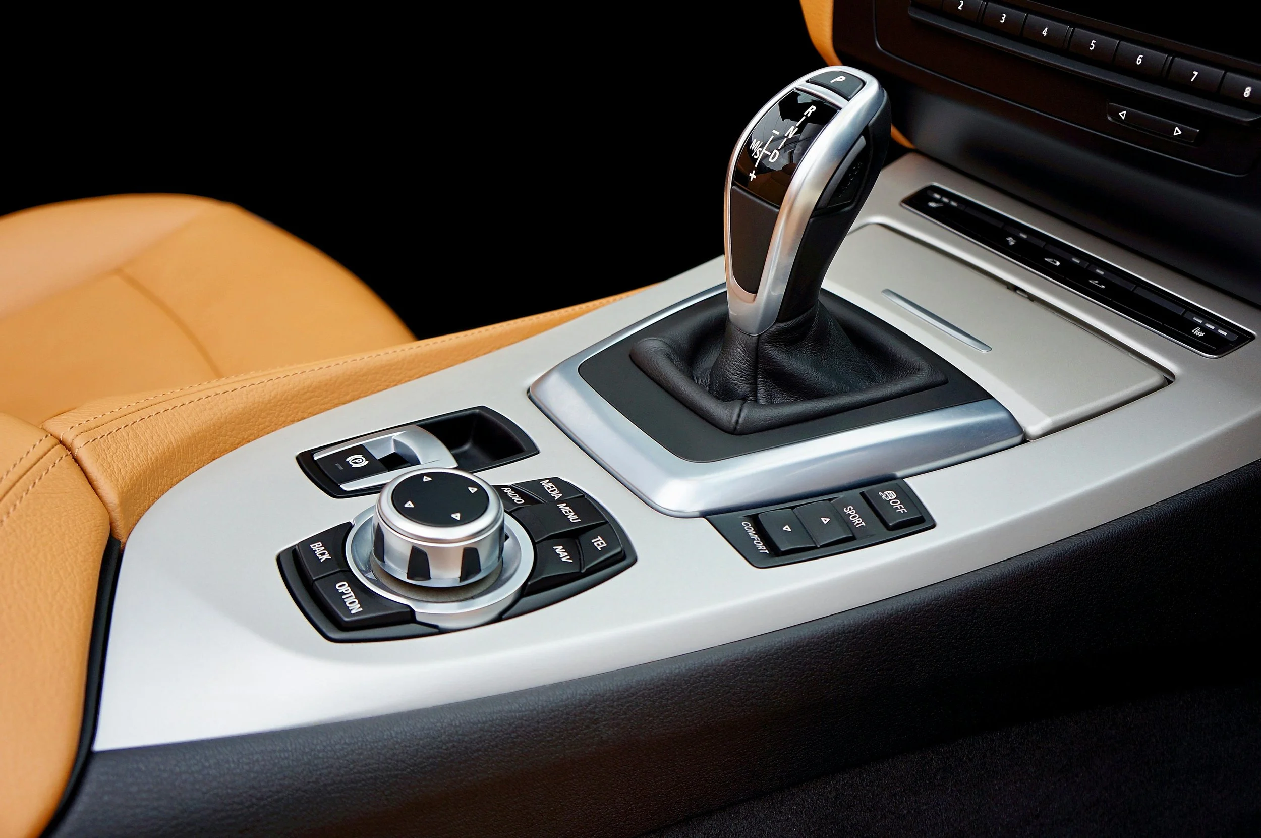 Close-up of a modern car's gear shift lever and control panel with buttons and a rotary dial, surrounded by beige leather interior.