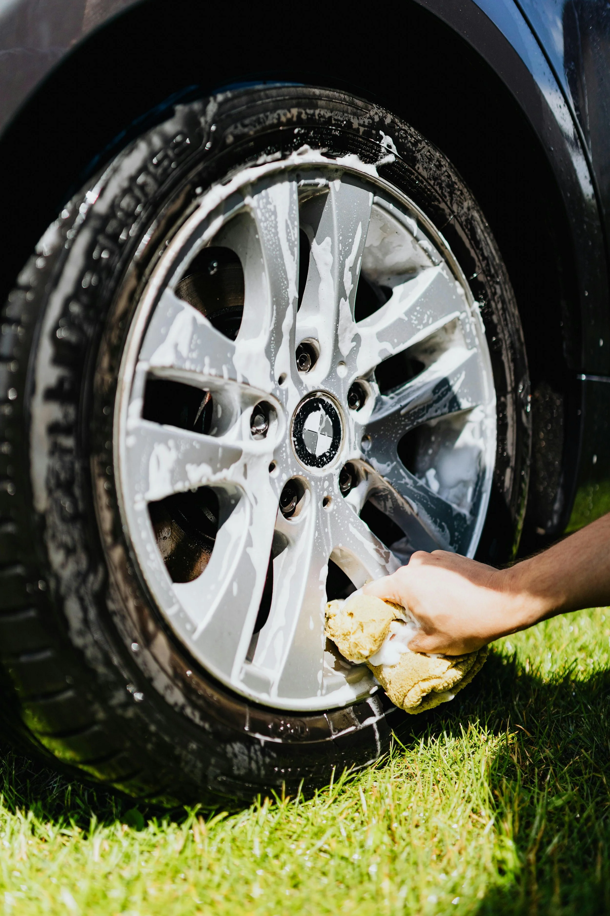 A person is cleaning a car wheel with soap and a sponge on grass.