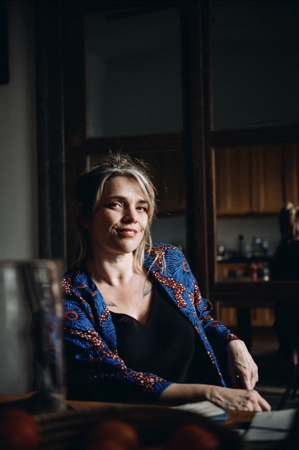 A woman with blonde hair sitting at a table in a dimly lit room, wearing a patterned blue and red jacket over a black top, smiling slightly at the camera.