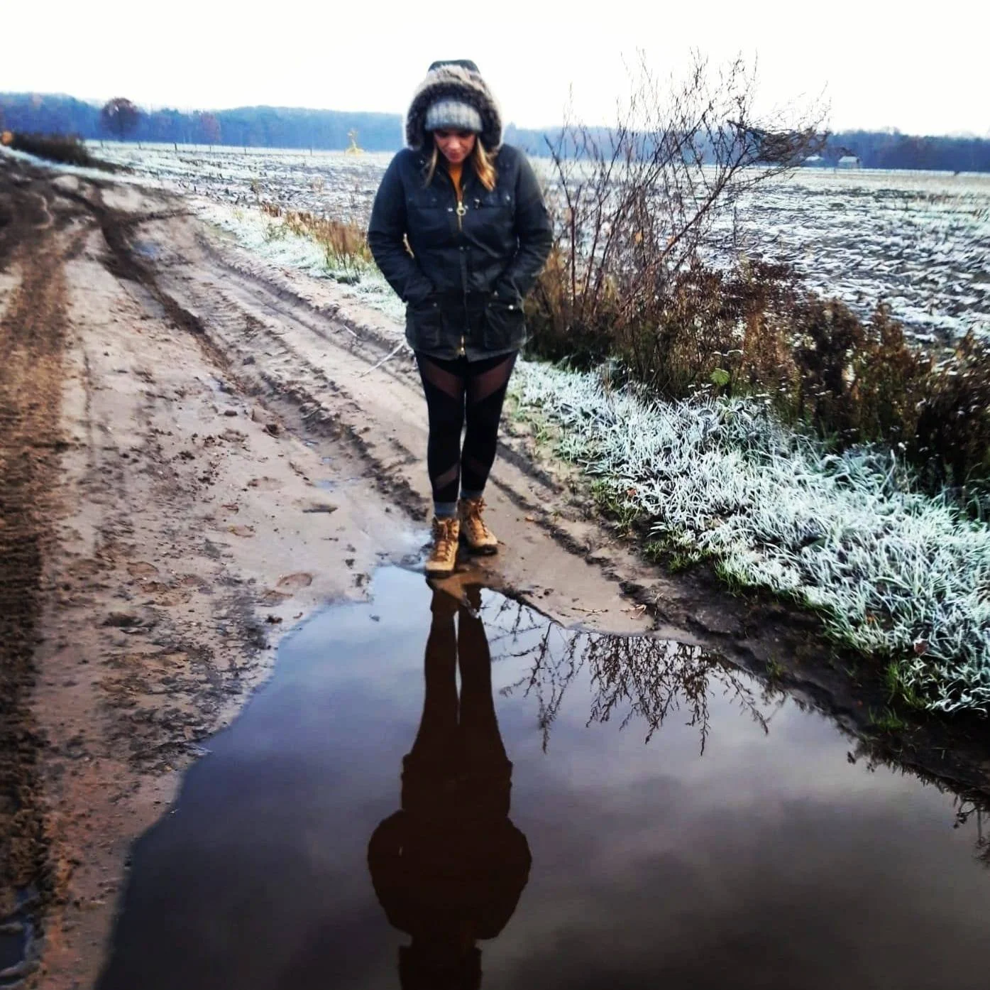 A woman in winter clothing, including a fur-lined hooded jacket, standing on a muddy dirt road looking at her reflection in a large puddle, with a snowy field and leafless trees in the background.