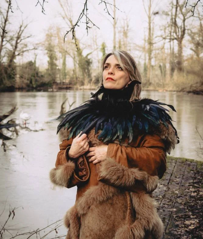 A woman standing by a river, wearing a brown faux fur coat and a feathered black cape, with trees in the background.